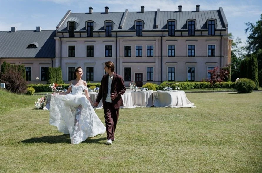 A bride and groom holding hands on a lawn in front of a large building with tables set for a wedding reception in the background.