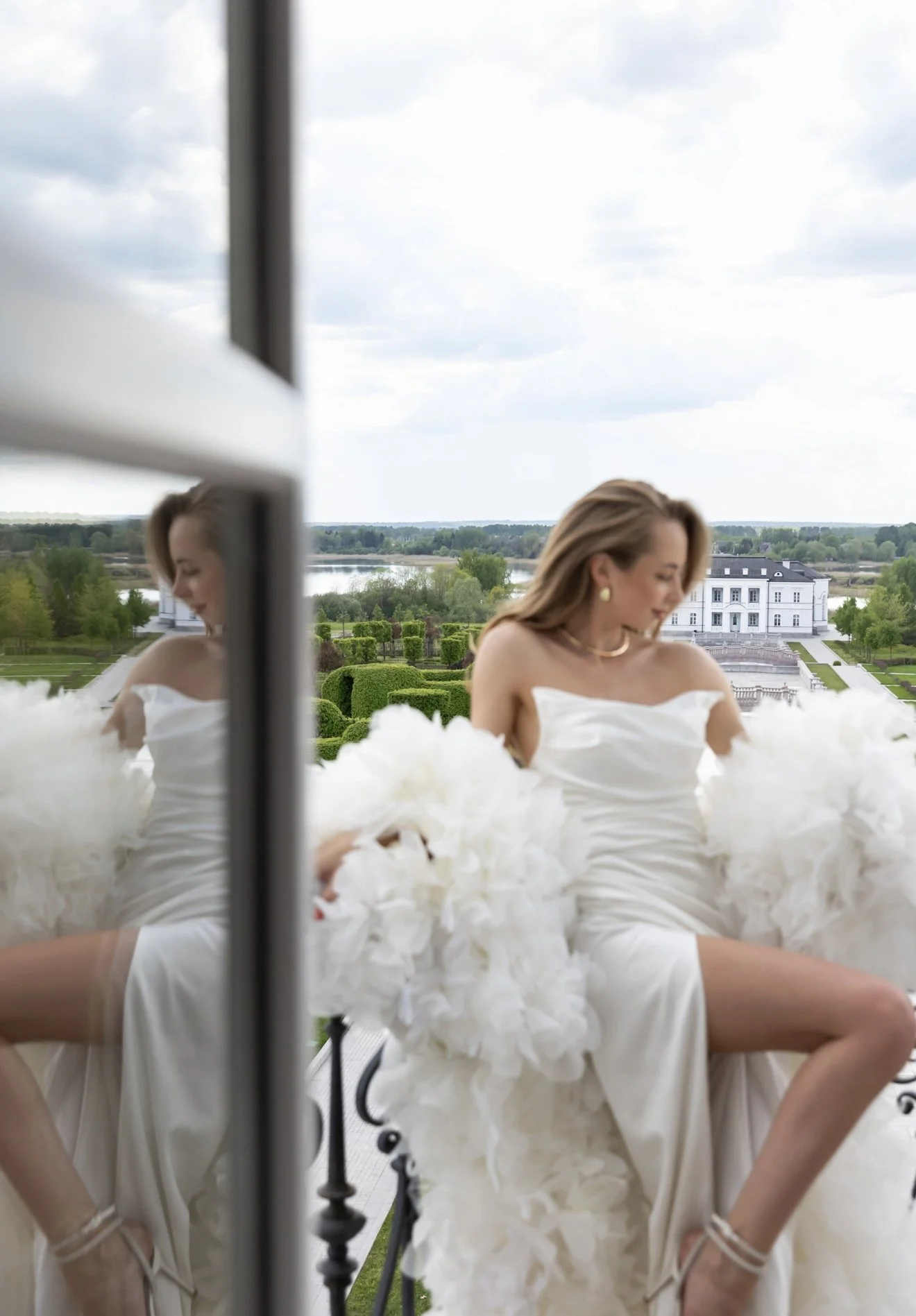 A woman in a white dress sitting outdoors on a balcony with a scenic landscape background, including a lake, trees, and a large white building, reflected in a mirror nearby.