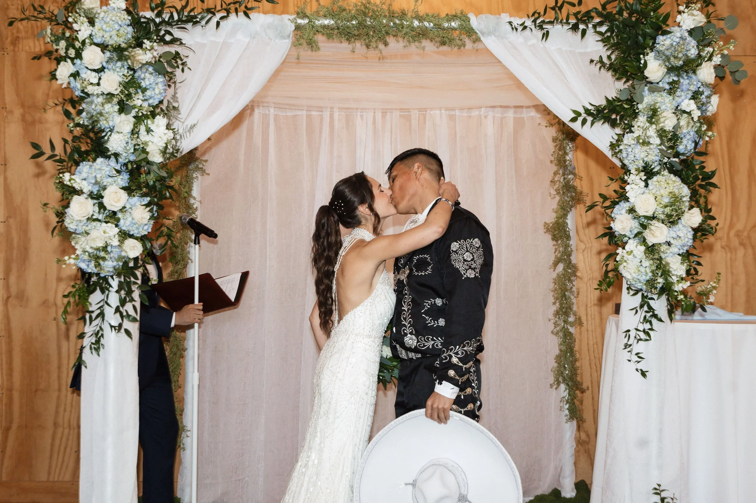 A bride and groom kiss during their wedding ceremony under a floral arch with white and blue flowers.