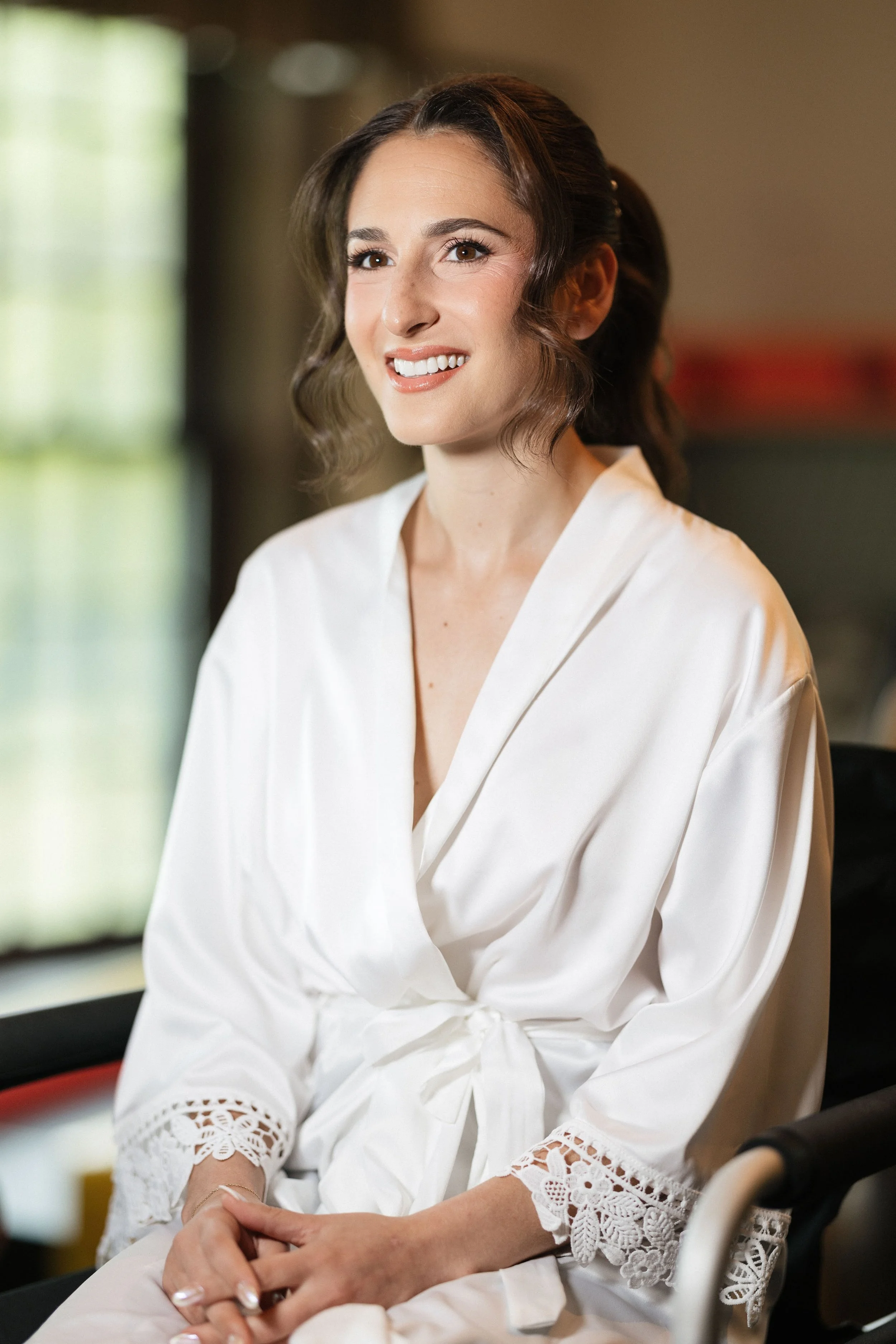 A woman with shoulder-length brown hair styled in soft waves, wearing a white blouse with embroidered lace cuffs, sitting indoors and smiling.