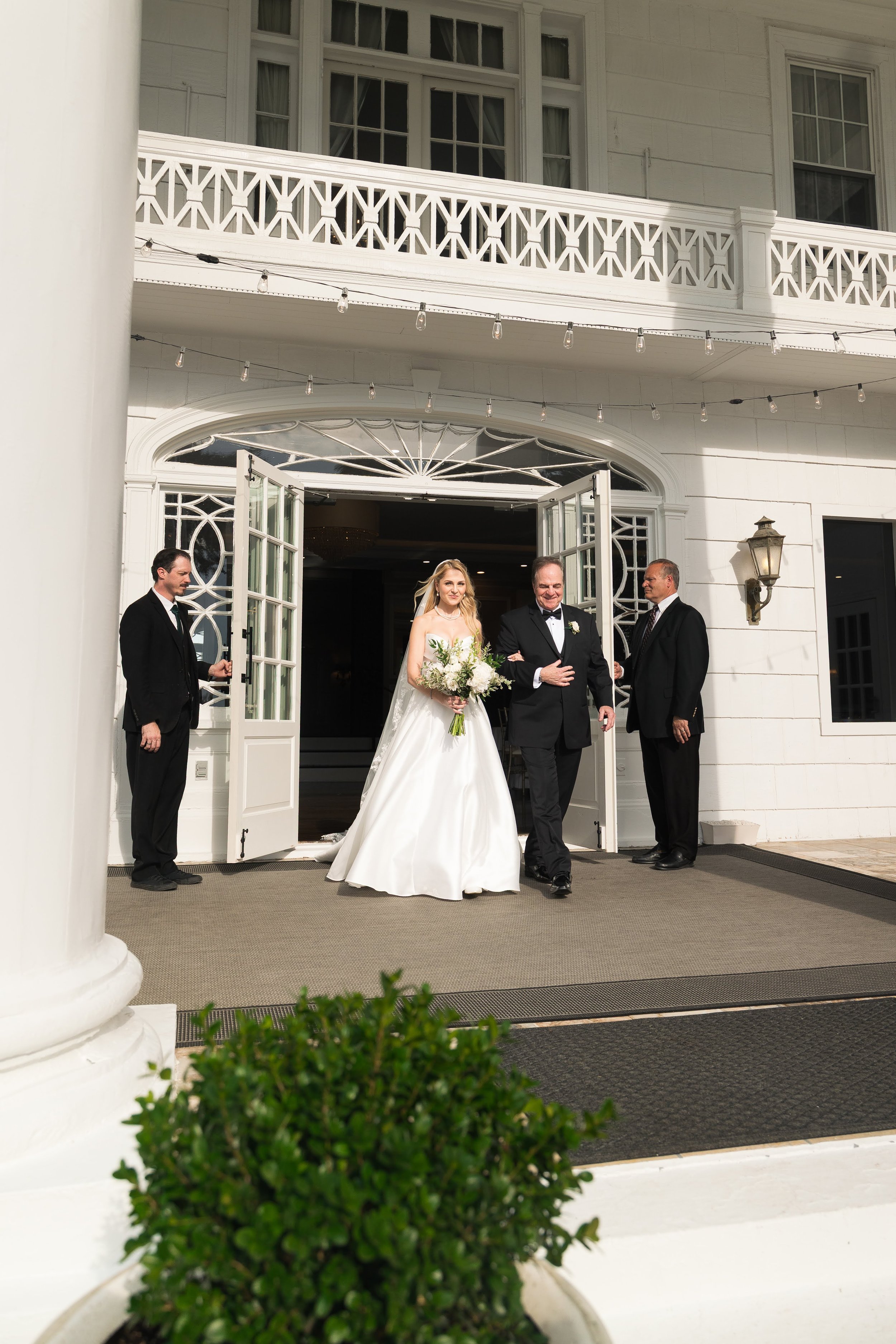 A bride and groom walking out of a white building with an elaborate door, accompanied by an older man, with two men in suits standing nearby. The bride holds a large bouquet, and all are smiling. The building features white walls, a balcony with deco