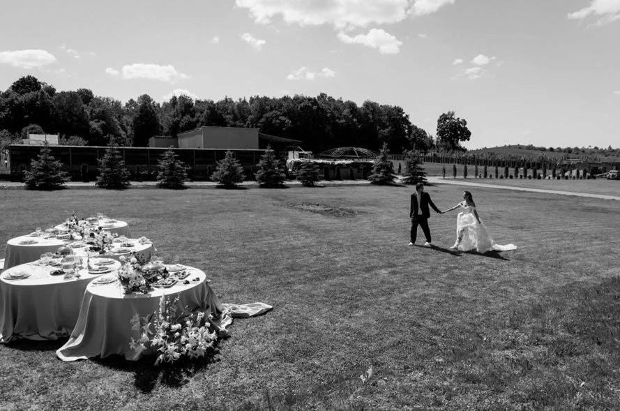 A couple dancing outdoors at a wedding reception with tables of food and flowers on the left, on a grassy area under a partly cloudy sky.