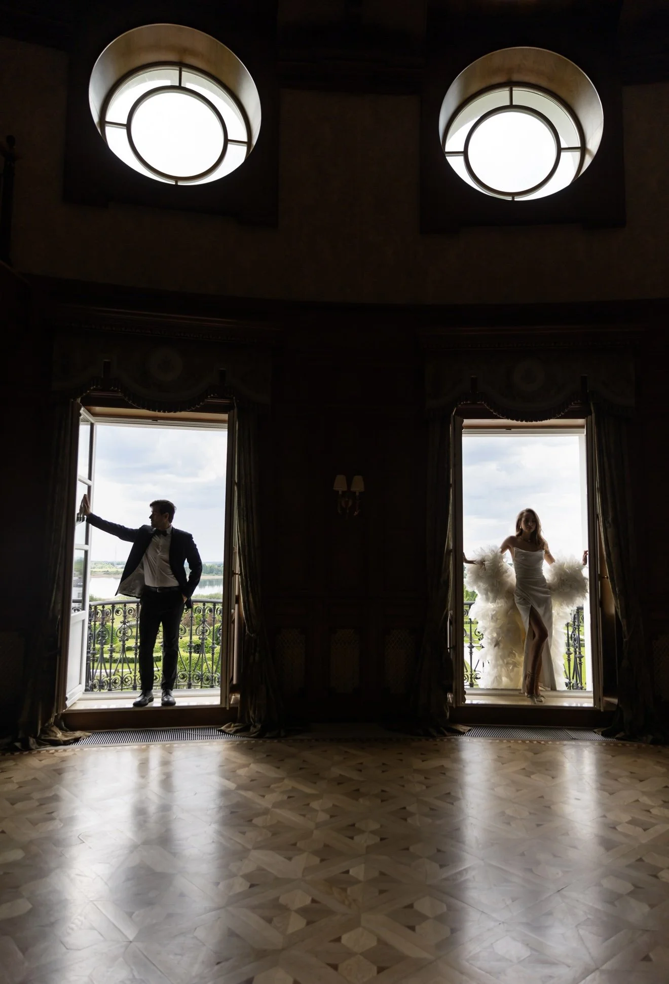 Two people standing on open balcony doors inside a grand room with ornate curtains. One is a man in a tuxedo, leaning against the door frame, and the other is a woman in a white dress with fluffy accents, standing on the balcony.