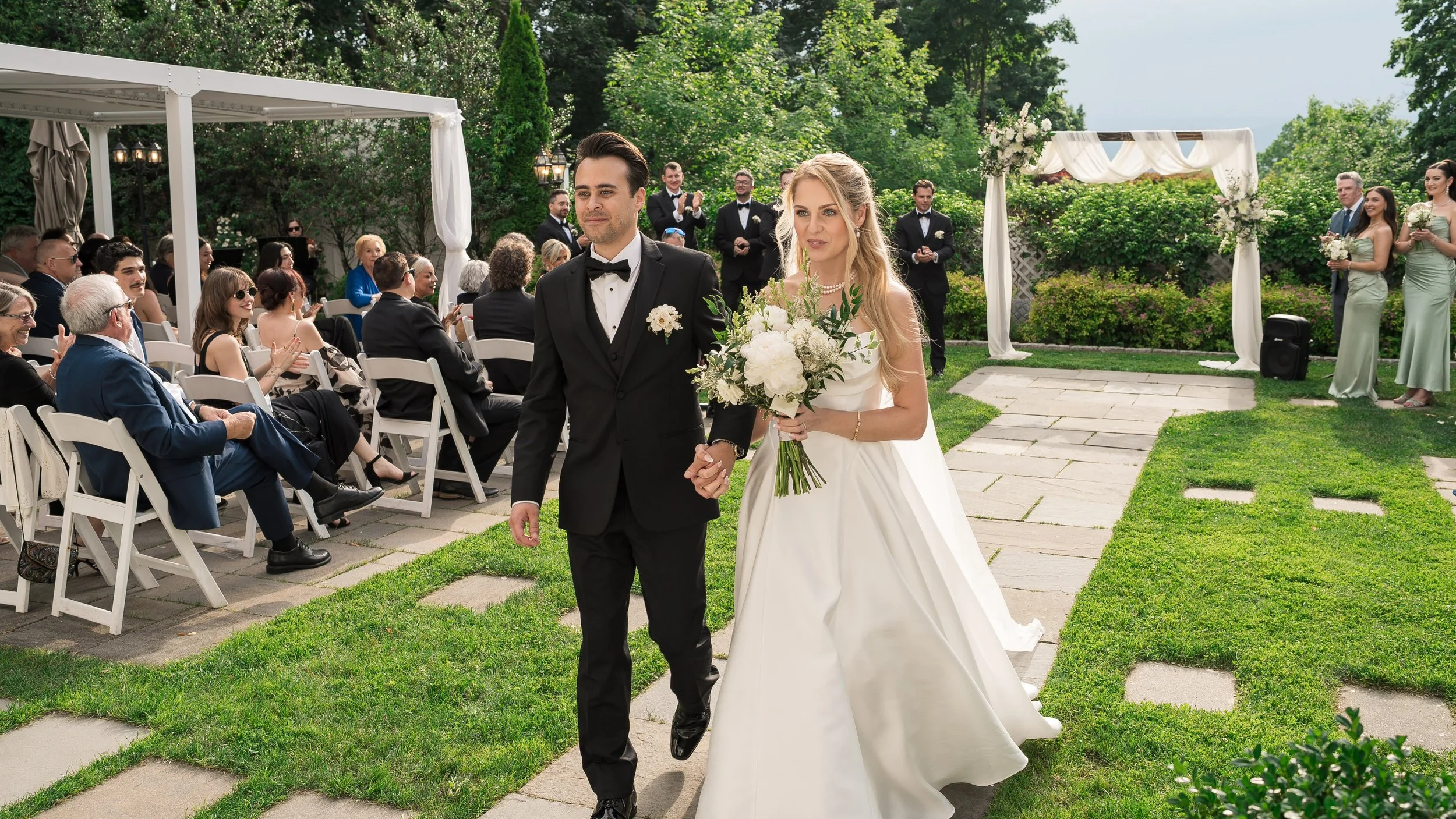 A bride and groom walk hand in hand down the aisle at an outdoor wedding ceremony, surrounded by seated guests and standing wedding party, with greenery and floral decorations in the background.