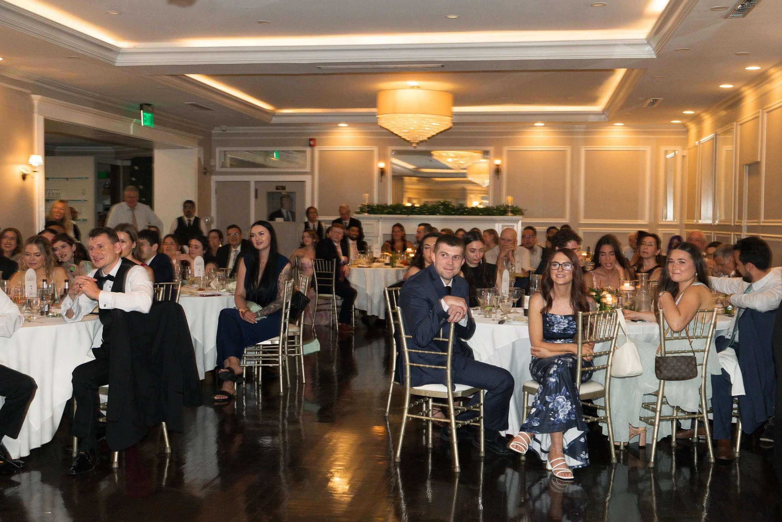 A large group of people dressed in formal attire sitting at decorated banquet tables in an elegant wedding reception hall with chandeliers.