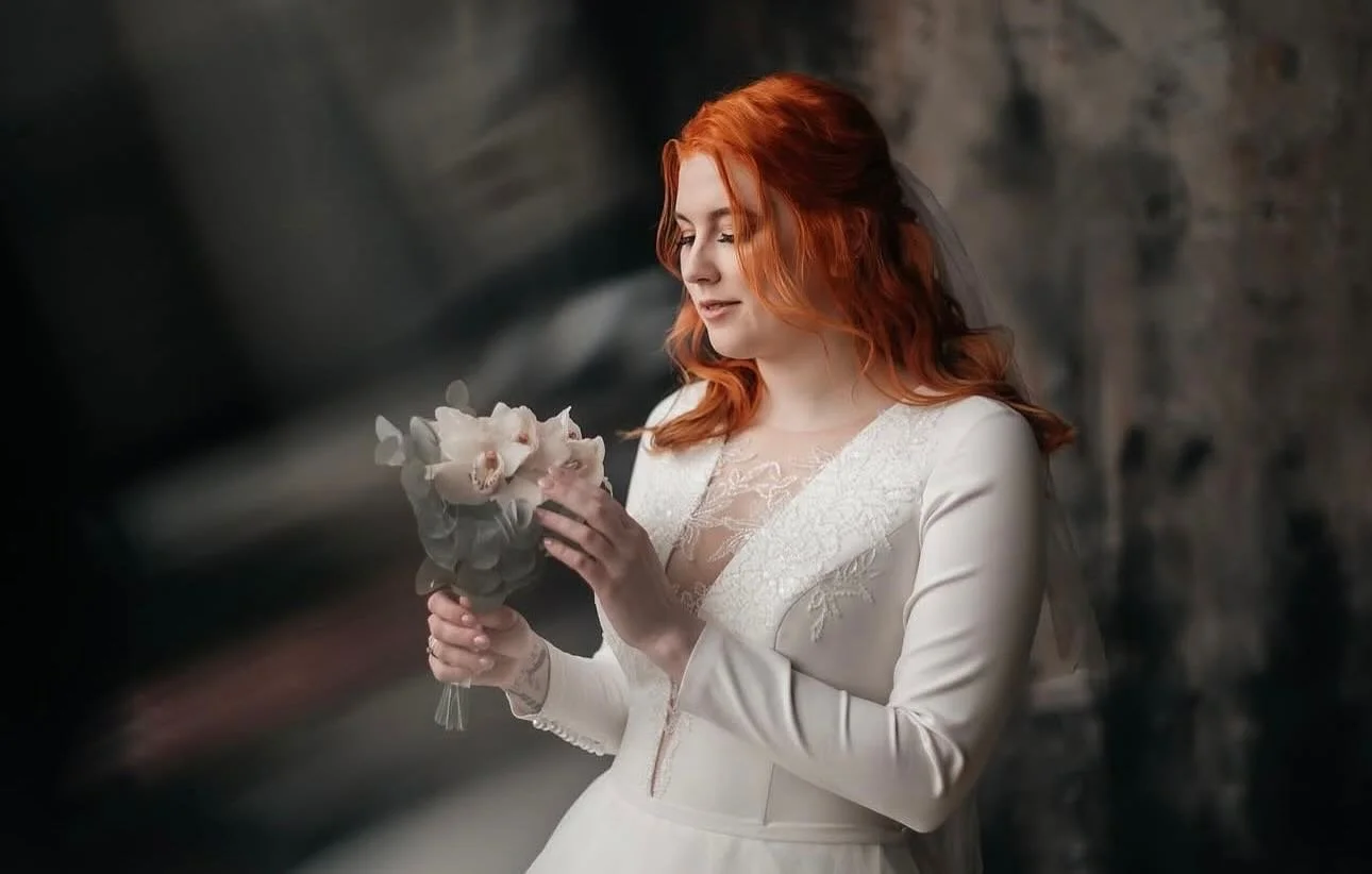 A woman with red hair in a white wedding dress holding a bouquet of pale pink and white flowers, standing against a stone wall.