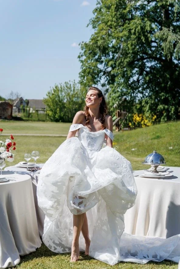 A woman in a white dress standing outdoors in a sunny setting with tables and chairs, trees, and a house in the background, smiling and enjoying the moment.