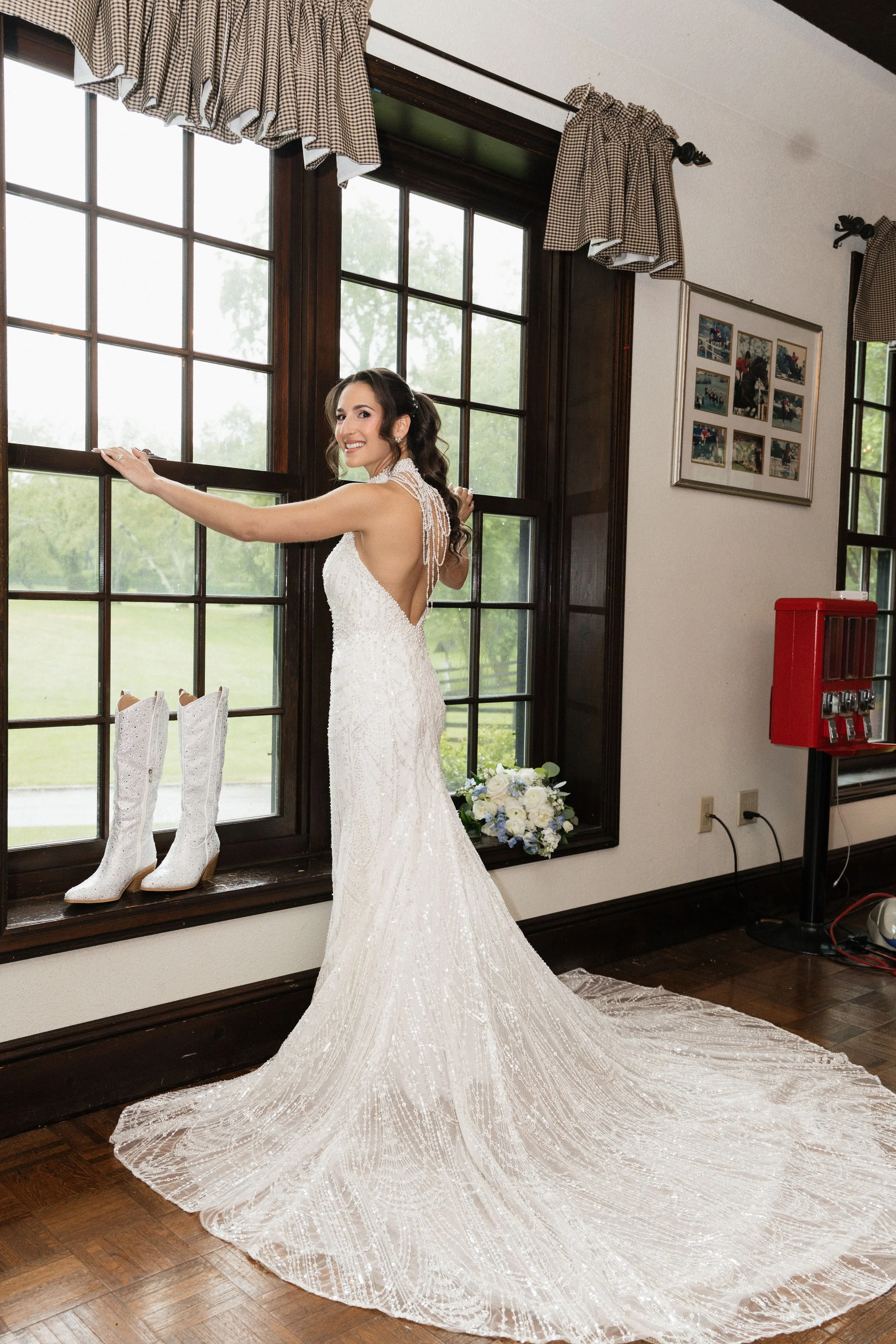 A bride in a white wedding gown with a long train stands by a window, smiling and holding onto the window frame. Two white cowboy boots are placed on the window sill, and a bouquet of white and blue flowers is on the floor nearby. The room has dark w