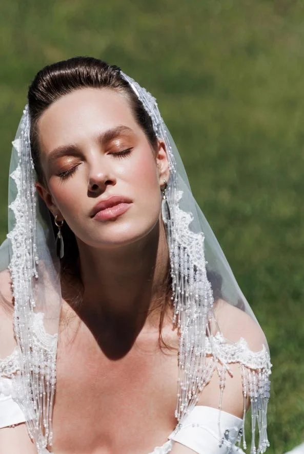 A woman with closed eyes, wearing a wedding veil and dress, standing outdoors on a grassy background.