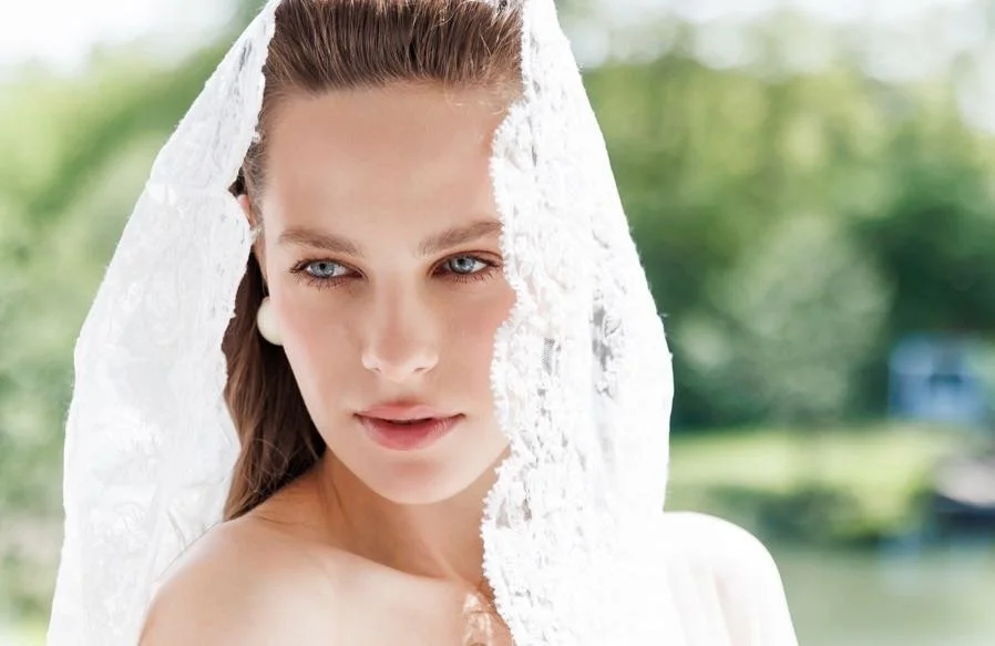 Close-up of a woman with light skin, blue eyes, and long brown hair, wearing a white lace veil, outdoors with a blurred green background.