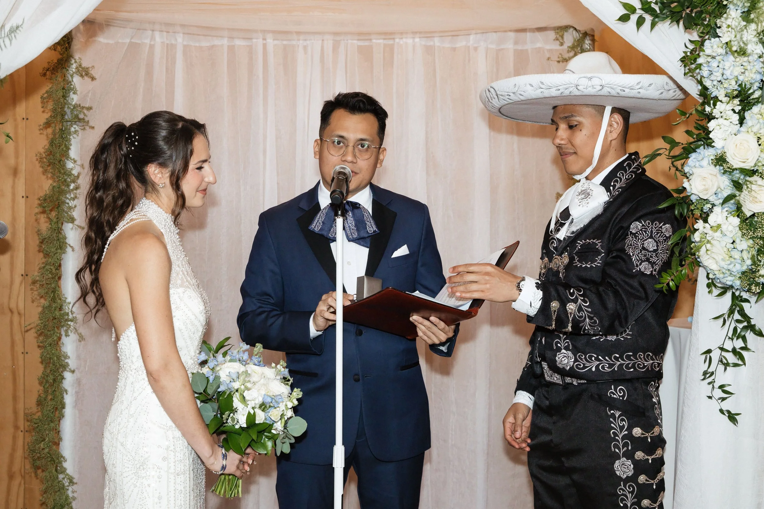 A wedding ceremony with a bride and groom standing before an officiant, exchanging vows, with floral decorations and a wooden backdrop.