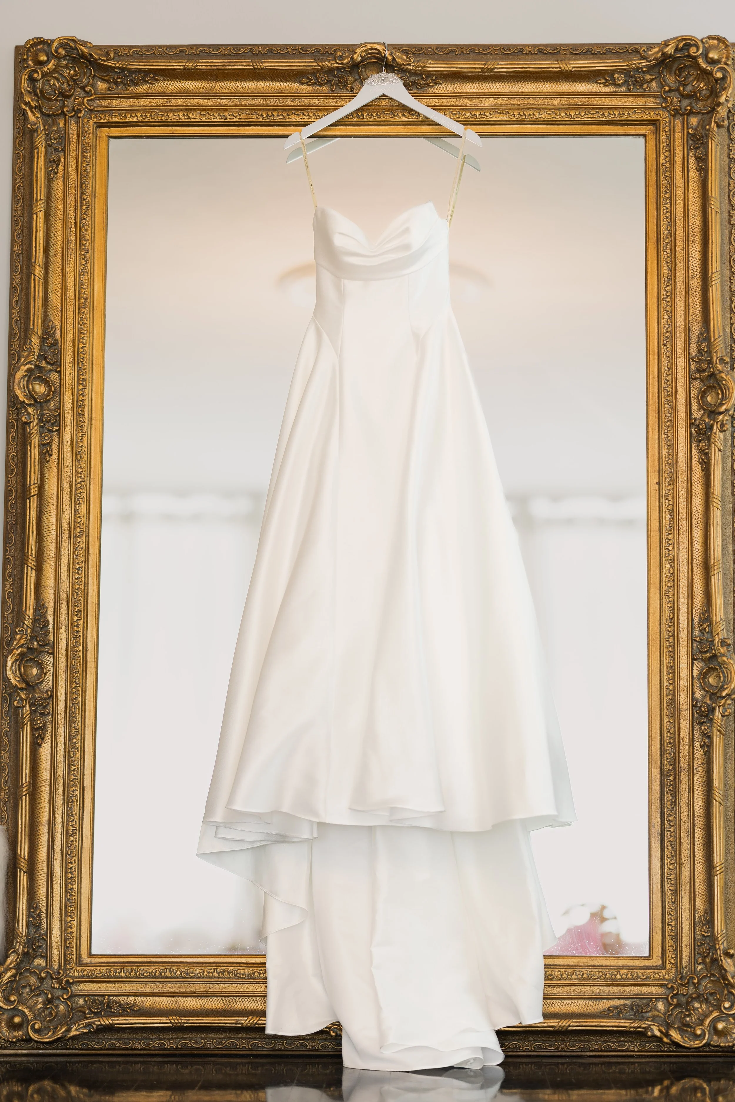 A white wedding dress hangs on a white hanger inside an ornate gold picture frame, which is positioned against a light background.
