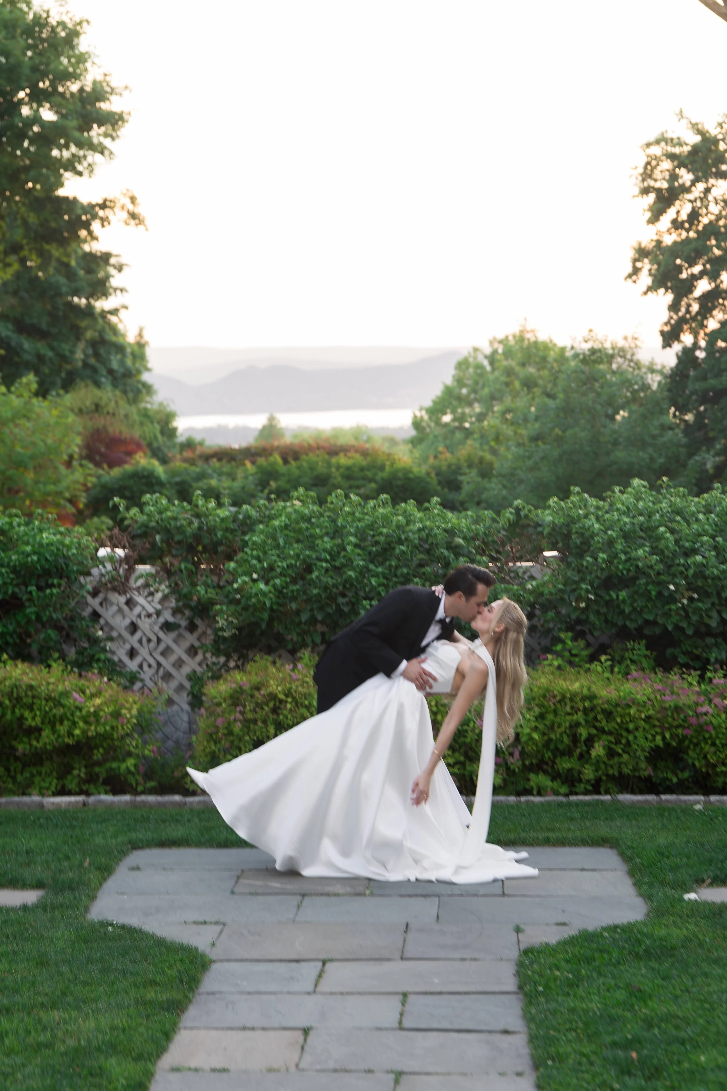 A newlywed couple in wedding attire sharing a kiss outdoors, with the groom dipping the bride while they kiss, surrounded by lush greenery and a scenic background.