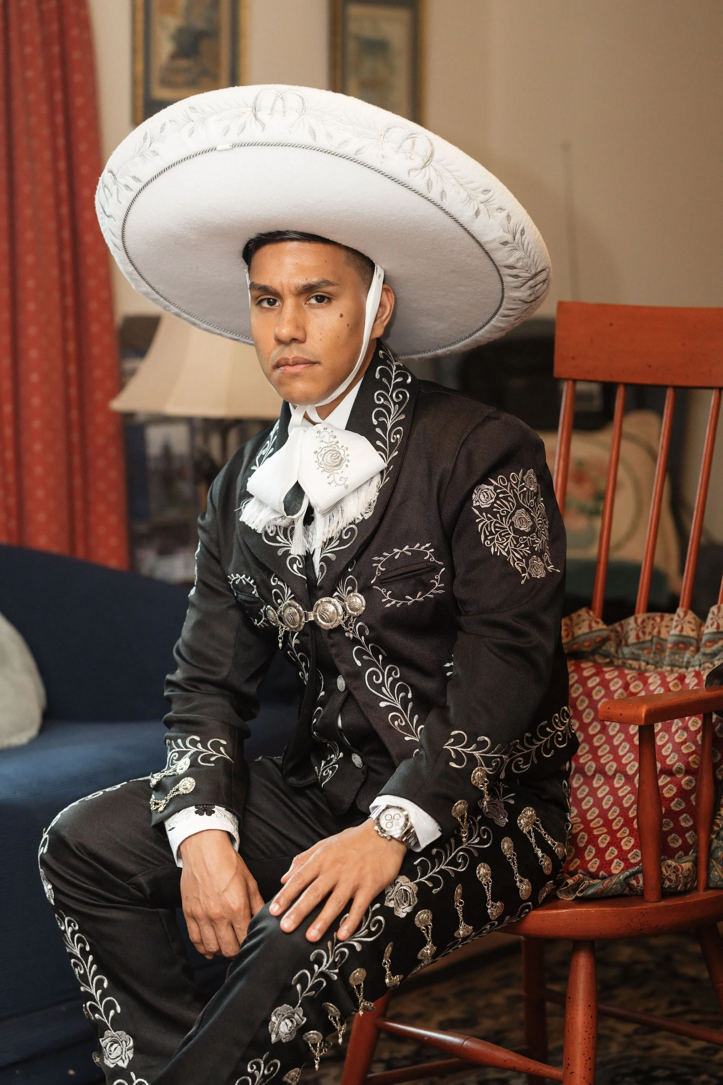 A man wearing a traditional Mexican charro suit, black with white embroidery and silver adornments, sitting on a wooden chair. He has a large white sombrero with embroidery and a white chinstrap. The background features a living room with furniture, 