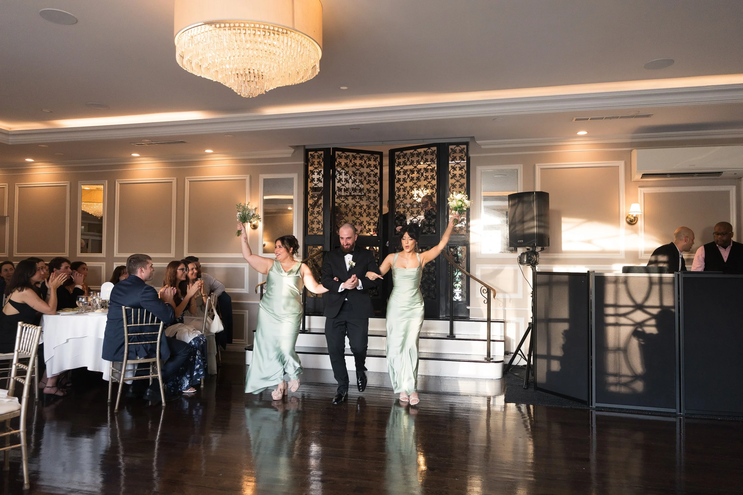 A bride and groom walking down the aisle of a wedding reception hall, smiling and holding hands, with two bridesmaids holding bouquets. Guests are clapping and watching them.