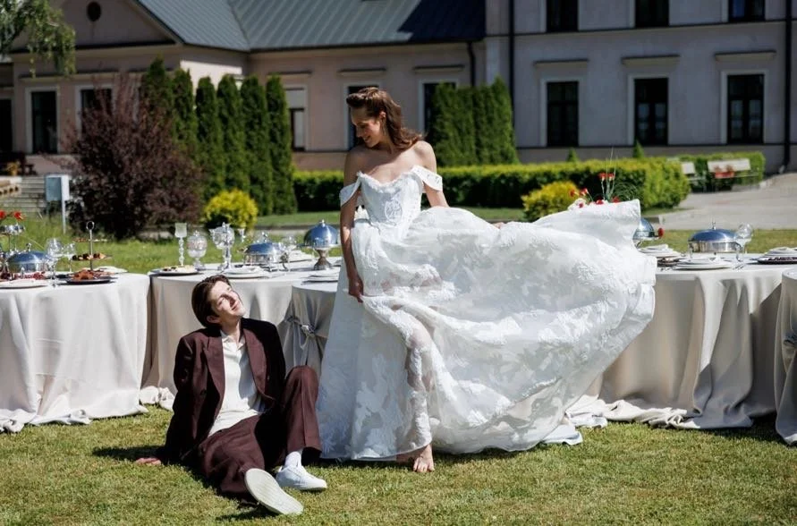 A woman in a white wedding dress playfully lifts her dress while standing on a grassy lawn, smiling at a man sitting on the ground in a brown suit during an outdoor wedding reception with tables set with silver serving dishes and glassware.
