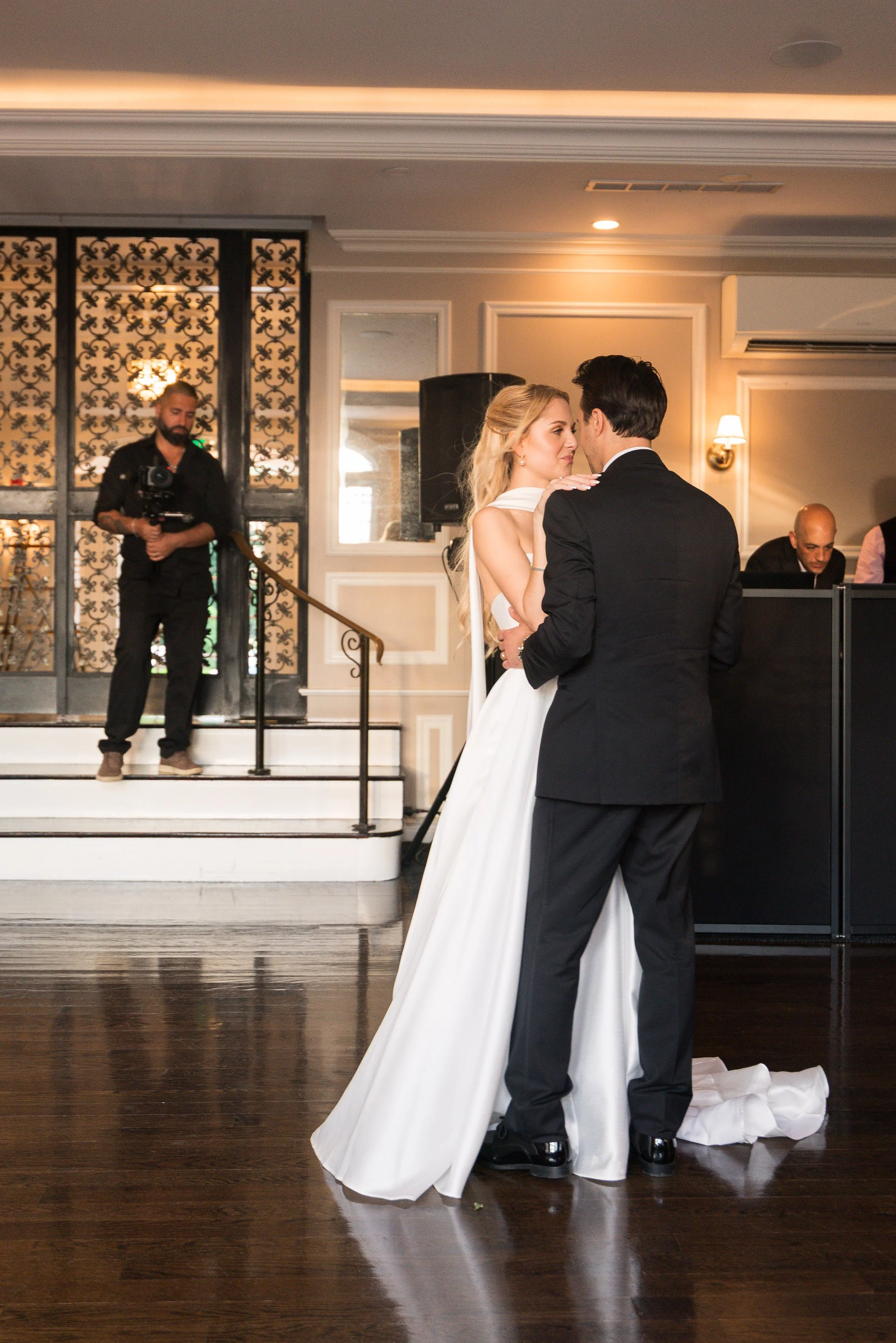 A bride and groom dance at their wedding reception, with the bride in a white gown and the groom in a black tuxedo, while a photographer and an event staff member observe nearby.
