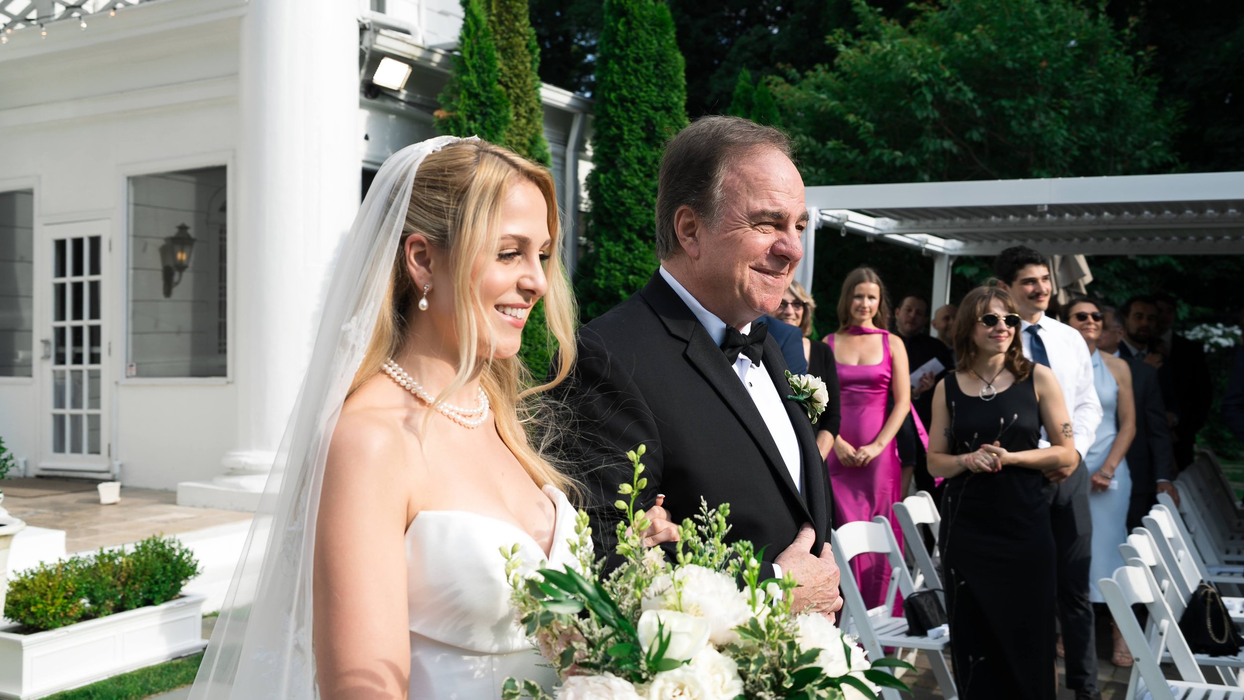 A bride and older man, possibly her father, standing at an outdoor wedding ceremony. The bride is smiling, wearing a white wedding dress, pearl earrings, and a veil. The man is dressed in a black tuxedo with a bowtie. Guests are visible in the backgr