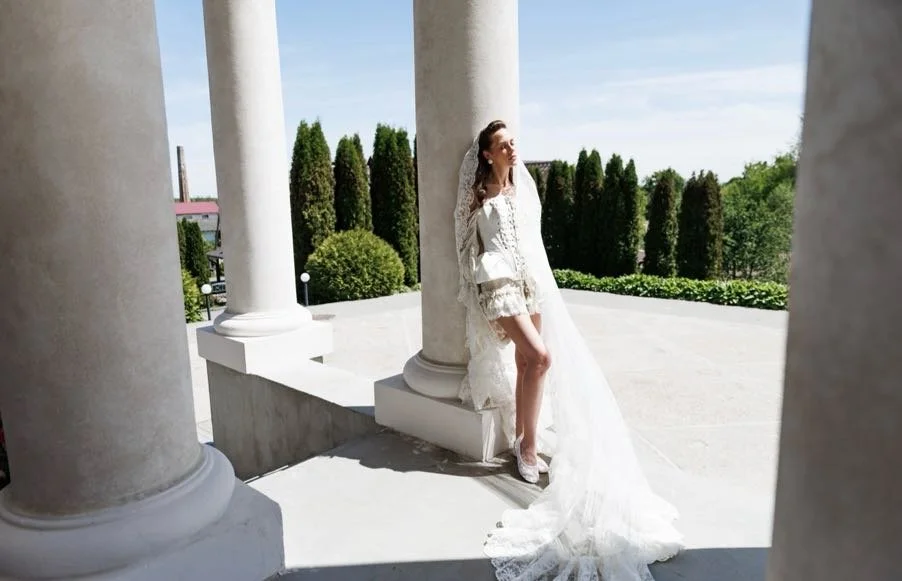 A woman in a white wedding or bridal gown with a long veil standing among large white classical columns outside, with green trees and blue sky in the background.