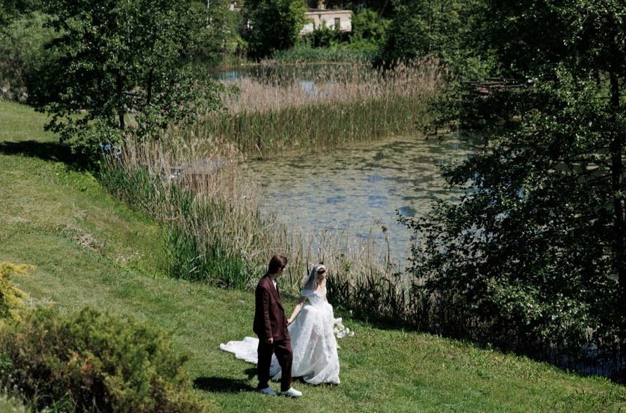 A bride and groom holding hands and walking near a pond in a lush green park.