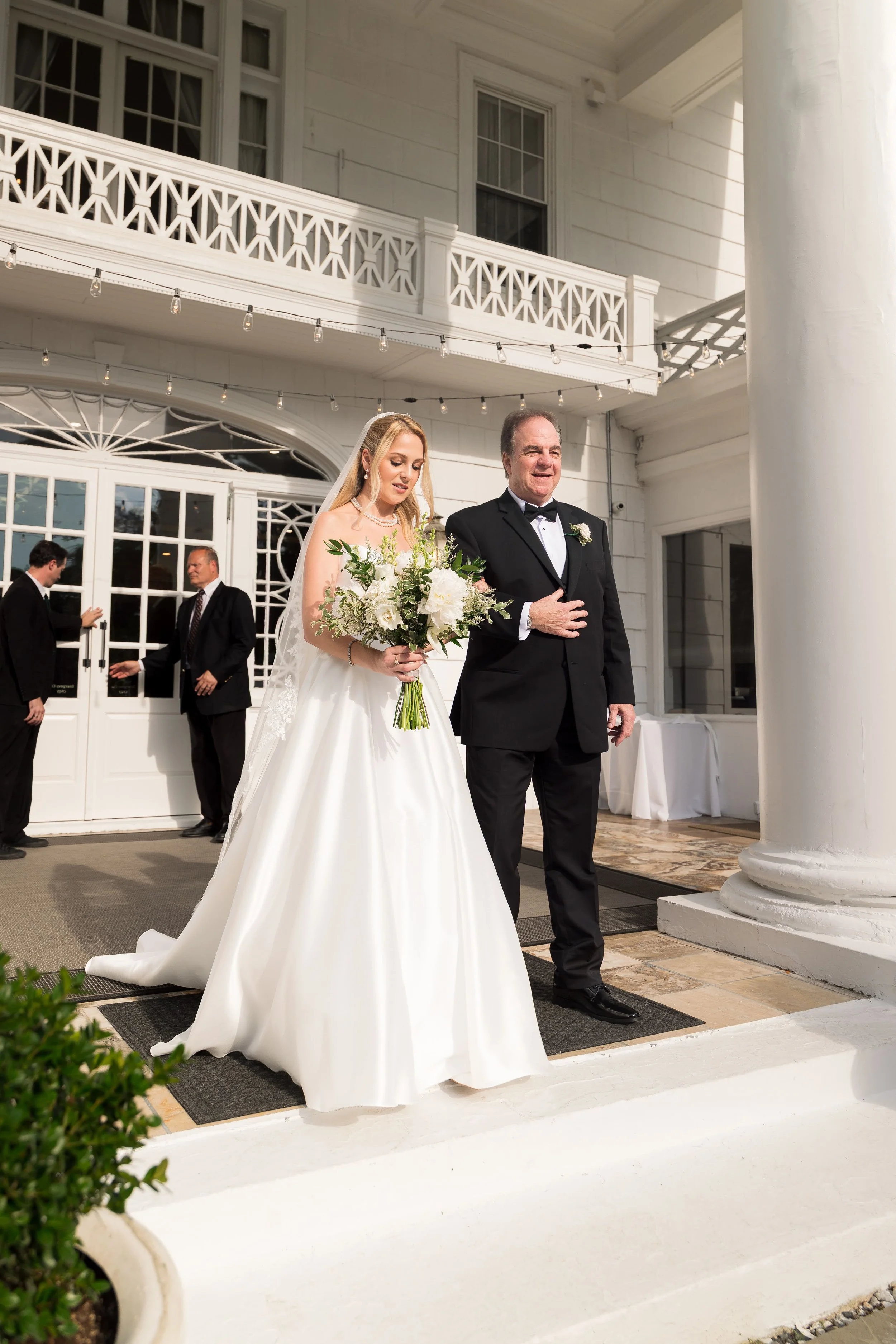 A bride in a white wedding gown holding a bouquet of white flowers walks arm-in-arm with a man in a black tuxedo outside a white colonial-style building during a wedding ceremony.