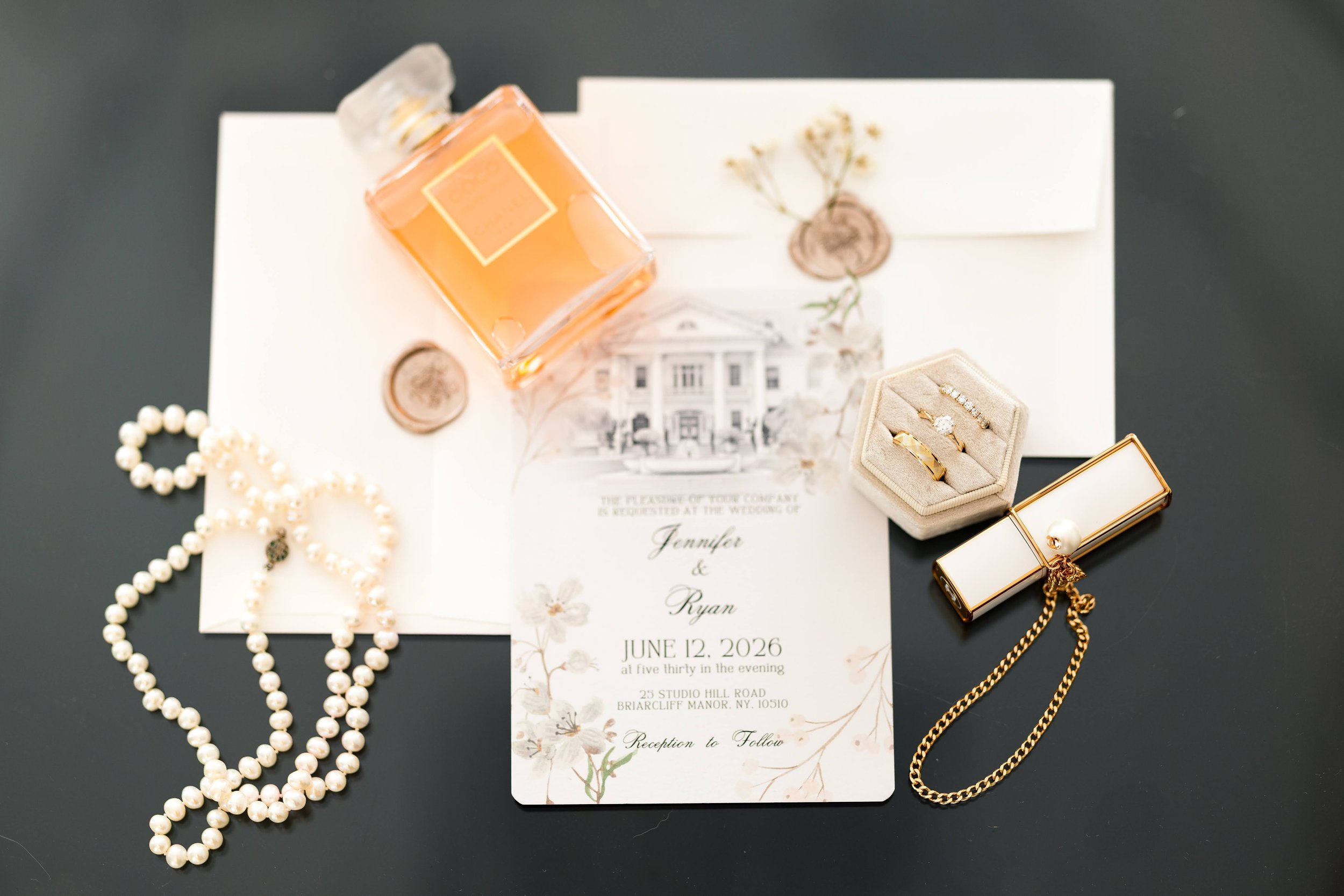 Wedding invitation surrounded by jewelry including gold rings, pearl necklace, and a pearl bracelet, with a bottle of perfume, on a dark surface.