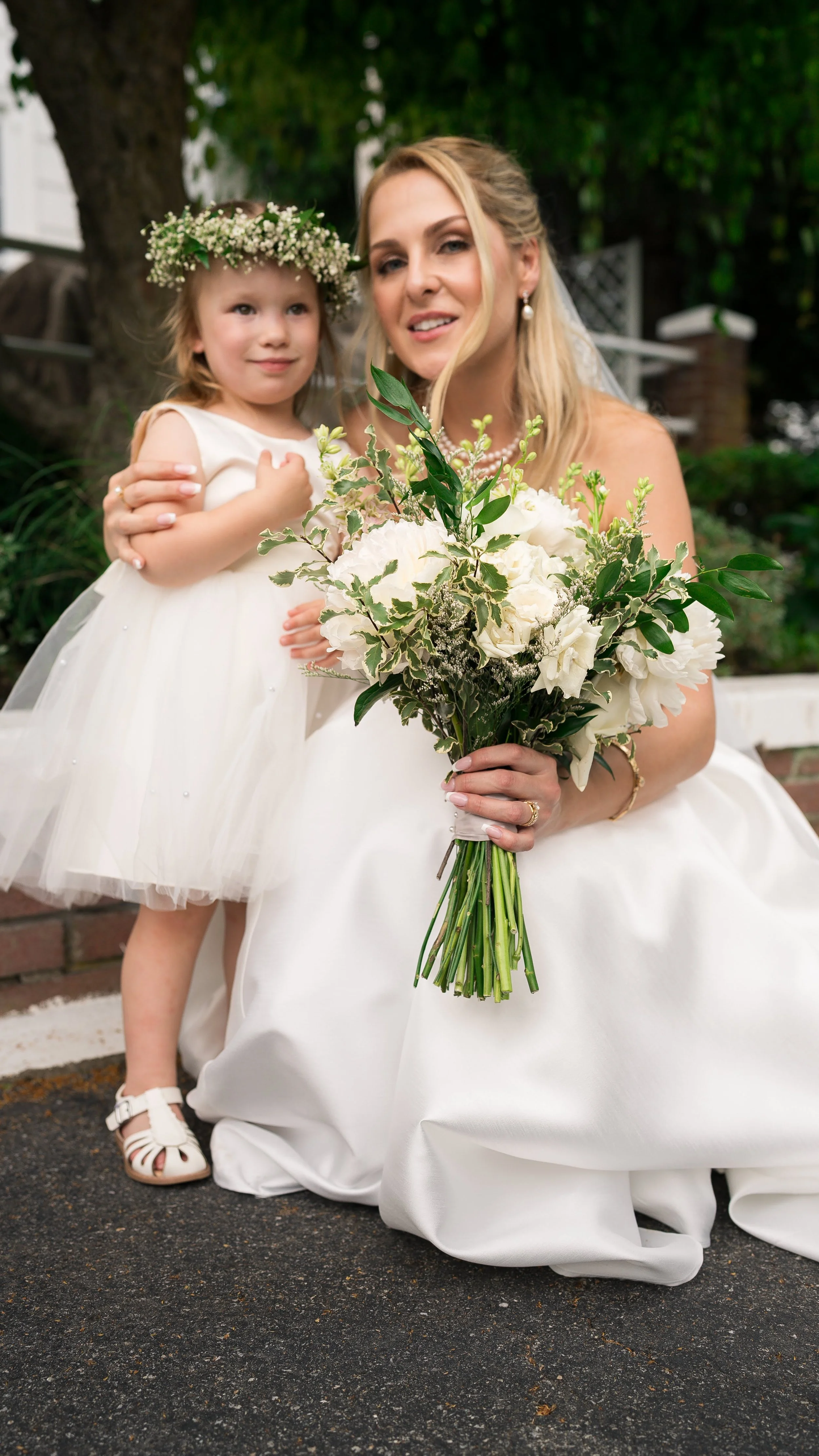 A woman in a wedding dress holding a bouquet of white flowers while sitting outdoors, with a young girl in a white dress and floral crown beside her, both smiling.