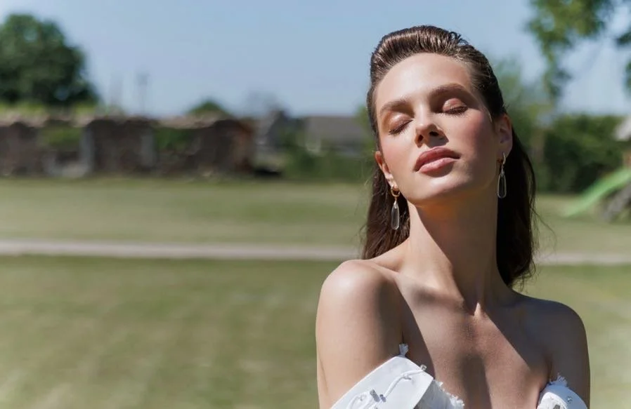 A woman with long brown hair and earrings stands outdoors with her eyes closed and face tilted upward, enjoying the sunlight.