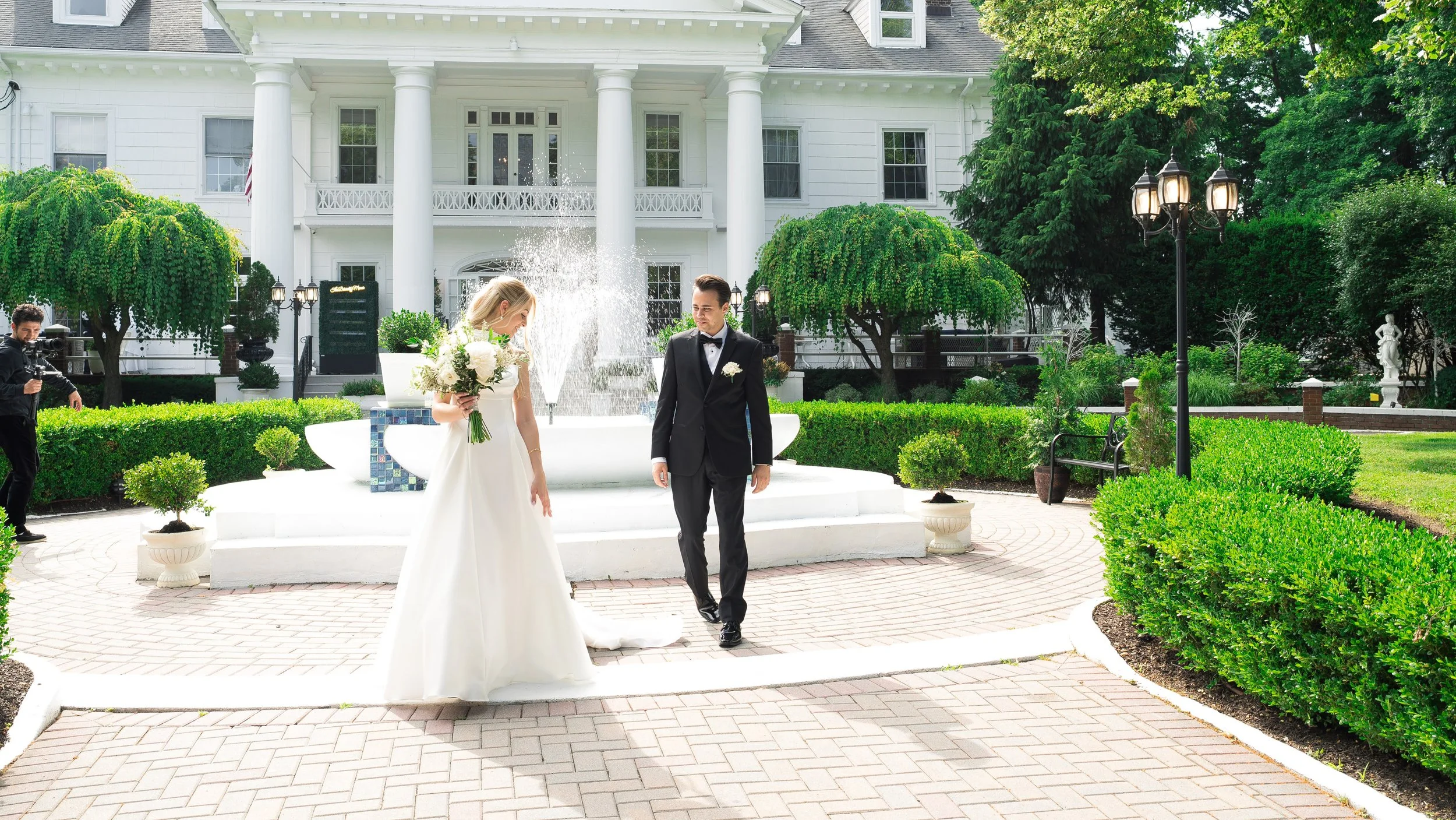 Couple getting married outdoors in front of a large white house with columns, surrounded by greenery, with a woman holding a bouquet and a man in a black tuxedo. A photographer is standing to the side.