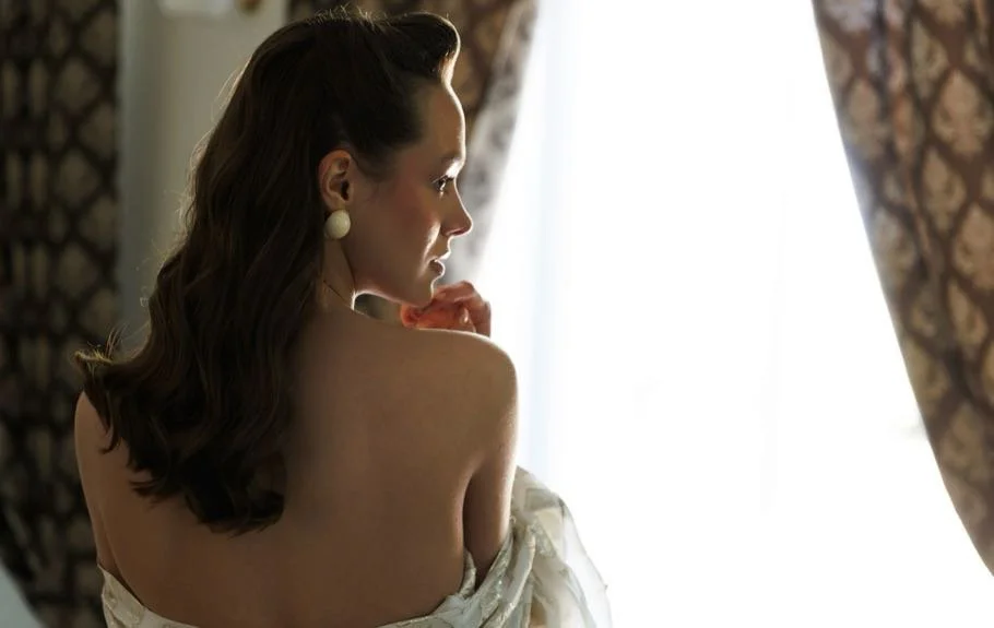 A woman with dark brown hair styled in loose waves, wearing large white earrings, gazes out a window with patterned curtains in the background.