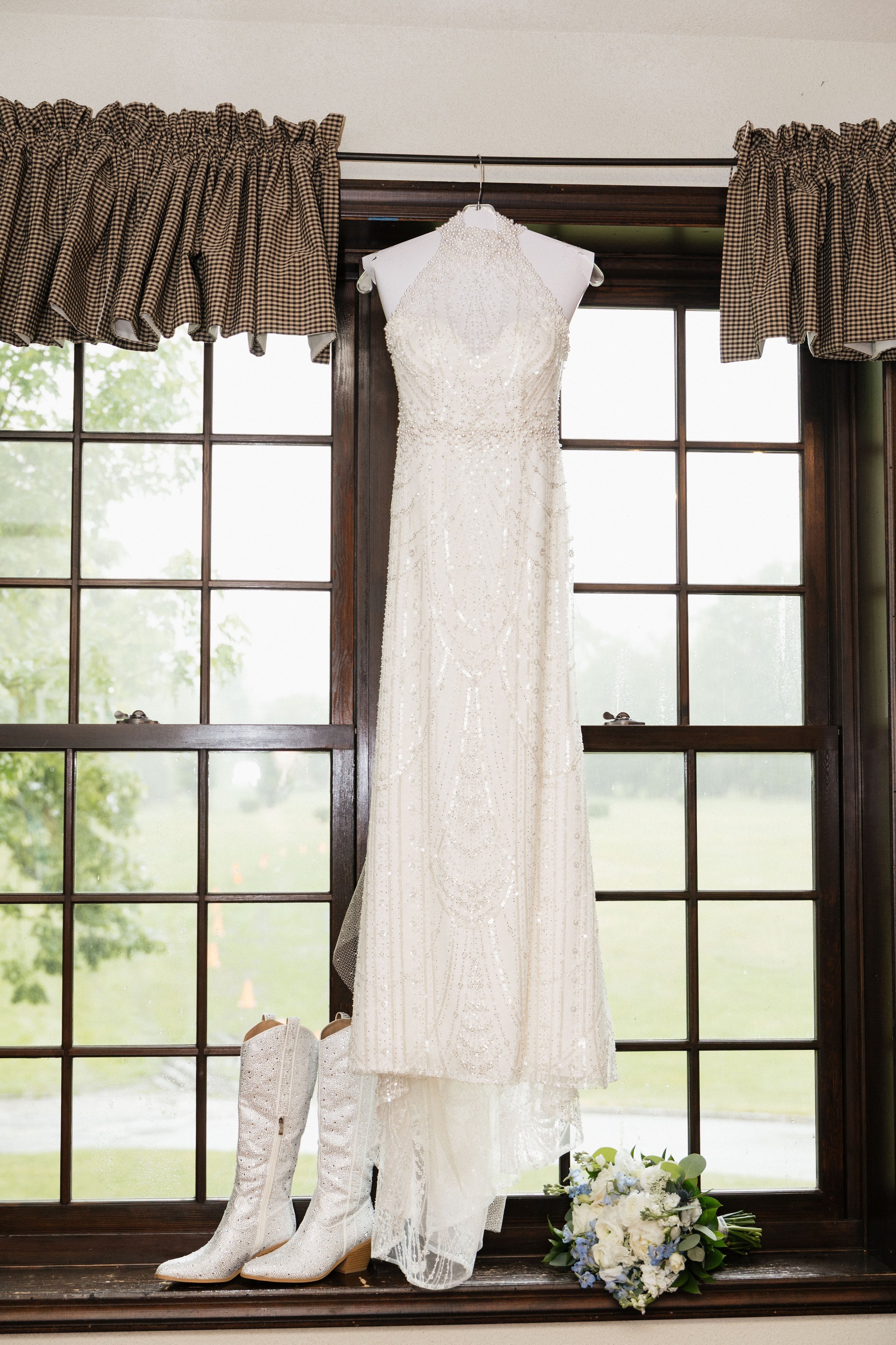 Wedding dress hanging in front of a window with curtains, white boots, bouquet of white and blue flowers on the floor.