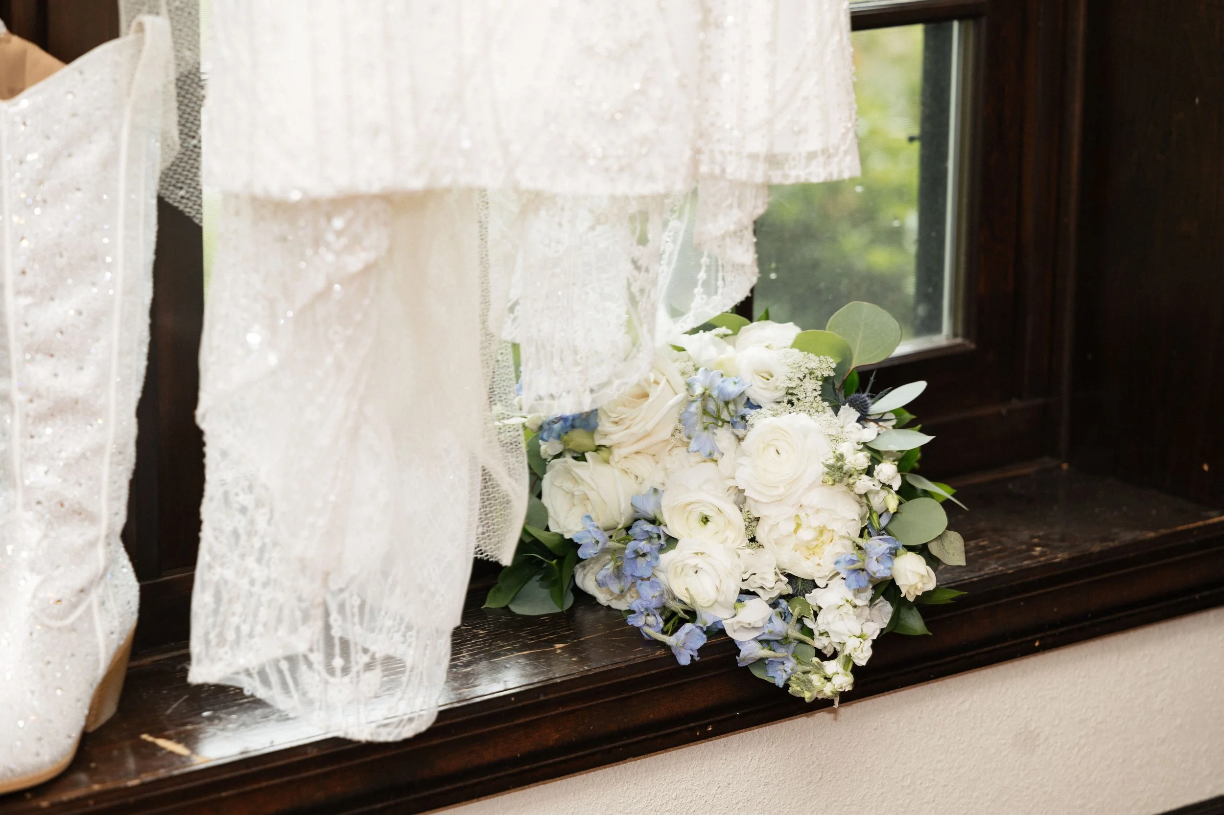 A bouquet of white and blue flowers resting on a dark wooden windowsill, with a lace curtain partially covering the window.