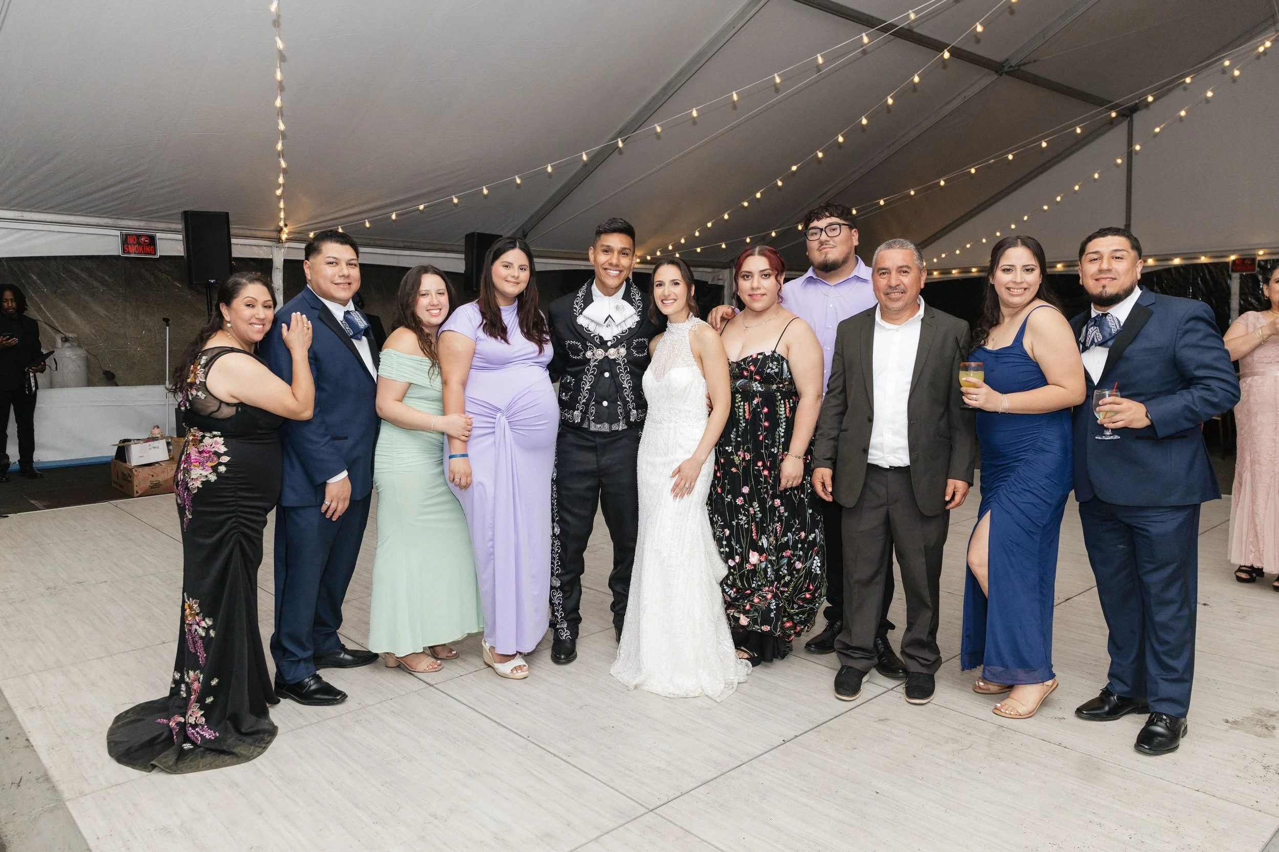 A group of people at a celebration, some dressed in formal attire including traditional Mexican clothing, standing under a canopy decorated with string lights, smiling for the photo.