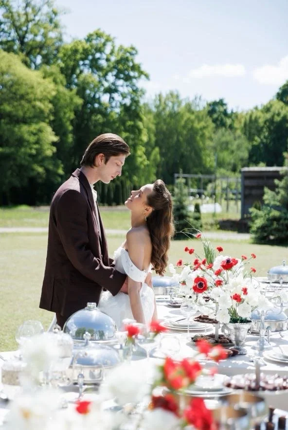 A young couple in wedding attire standing outdoors near a table with flowers and dishes, gazing at each other.
