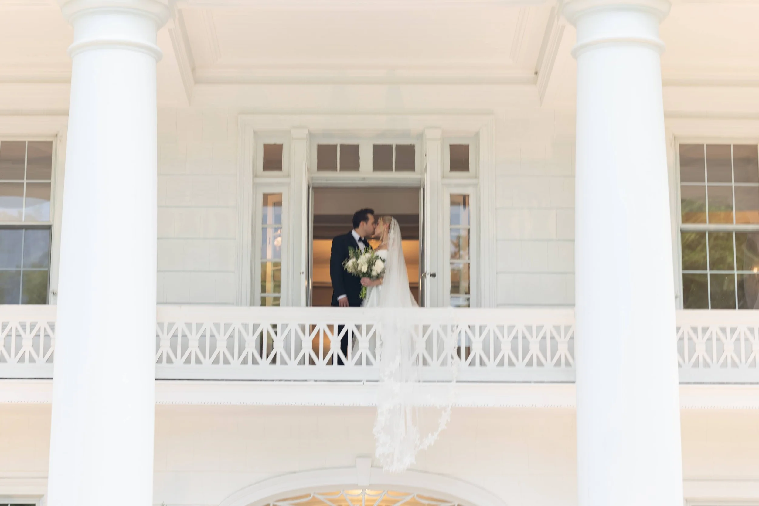 A bride and groom sharing a kiss on the porch of a white house with large pillars, windows, and a decorative white railing.