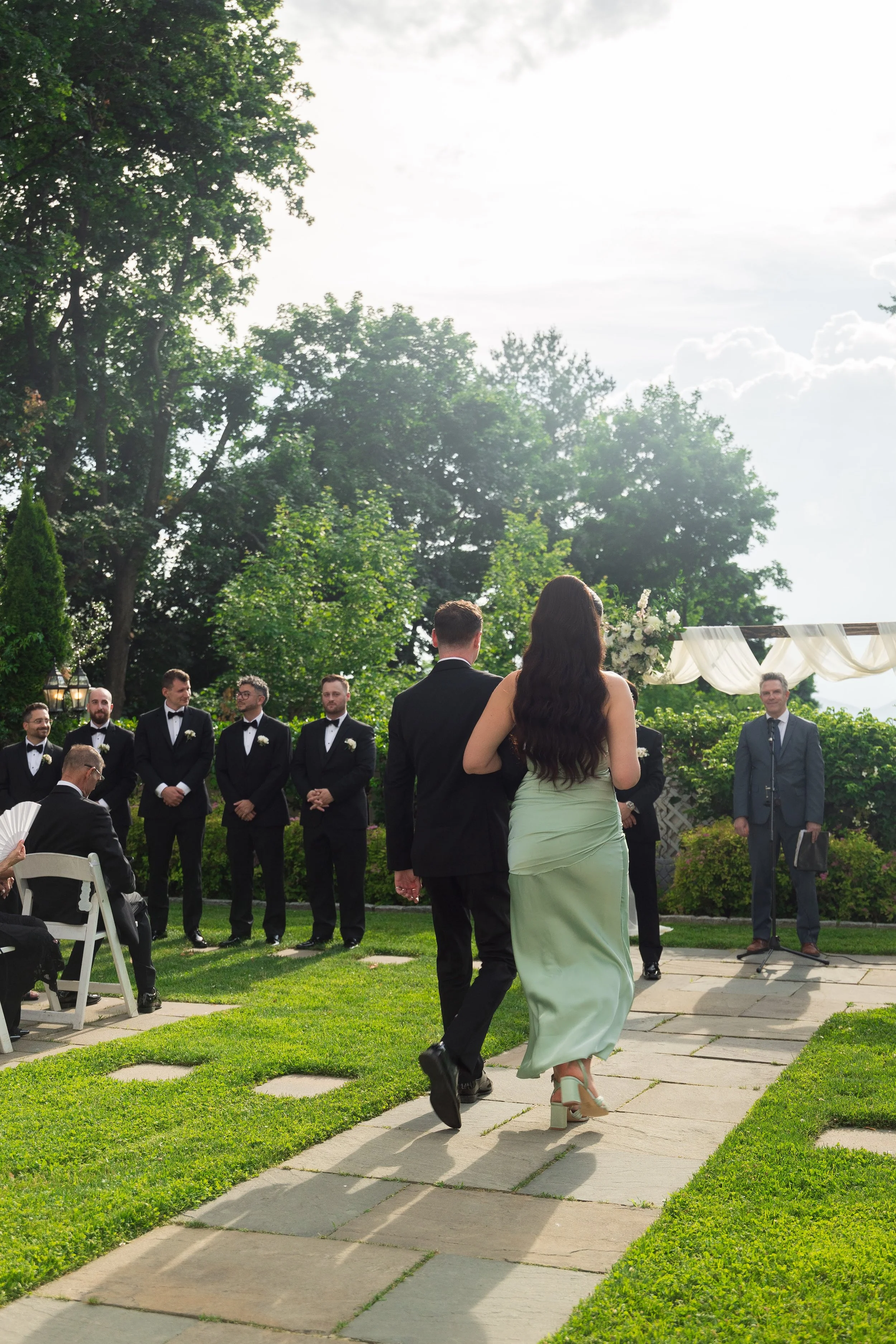 A wedding ceremony outdoors with a group of men in tuxedos standing in front of a draped white fabric arch, and a woman in a light green dress and high heels walking towards the group on a stone pathway. Green trees and a partly cloudy sky are in the