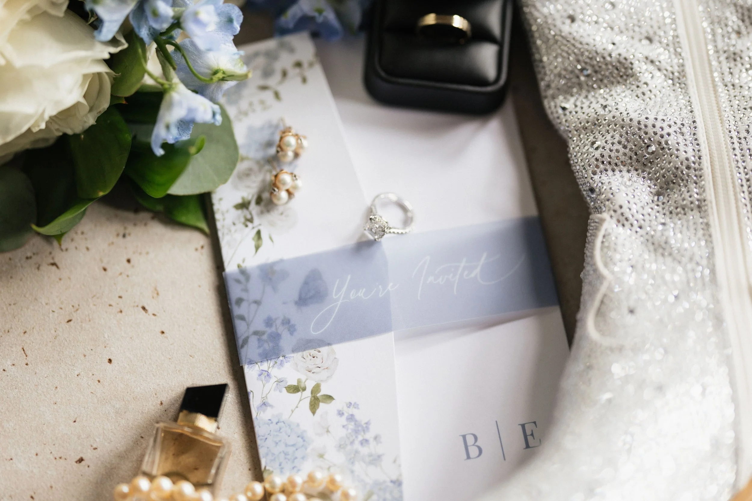 Close-up of jewelry, a ring with a large stone, a pair of pearl earrings, a pearl bracelet, and a floral-themed card, with a bouquet of white and blue flowers and a sparkly silver bag nearby.