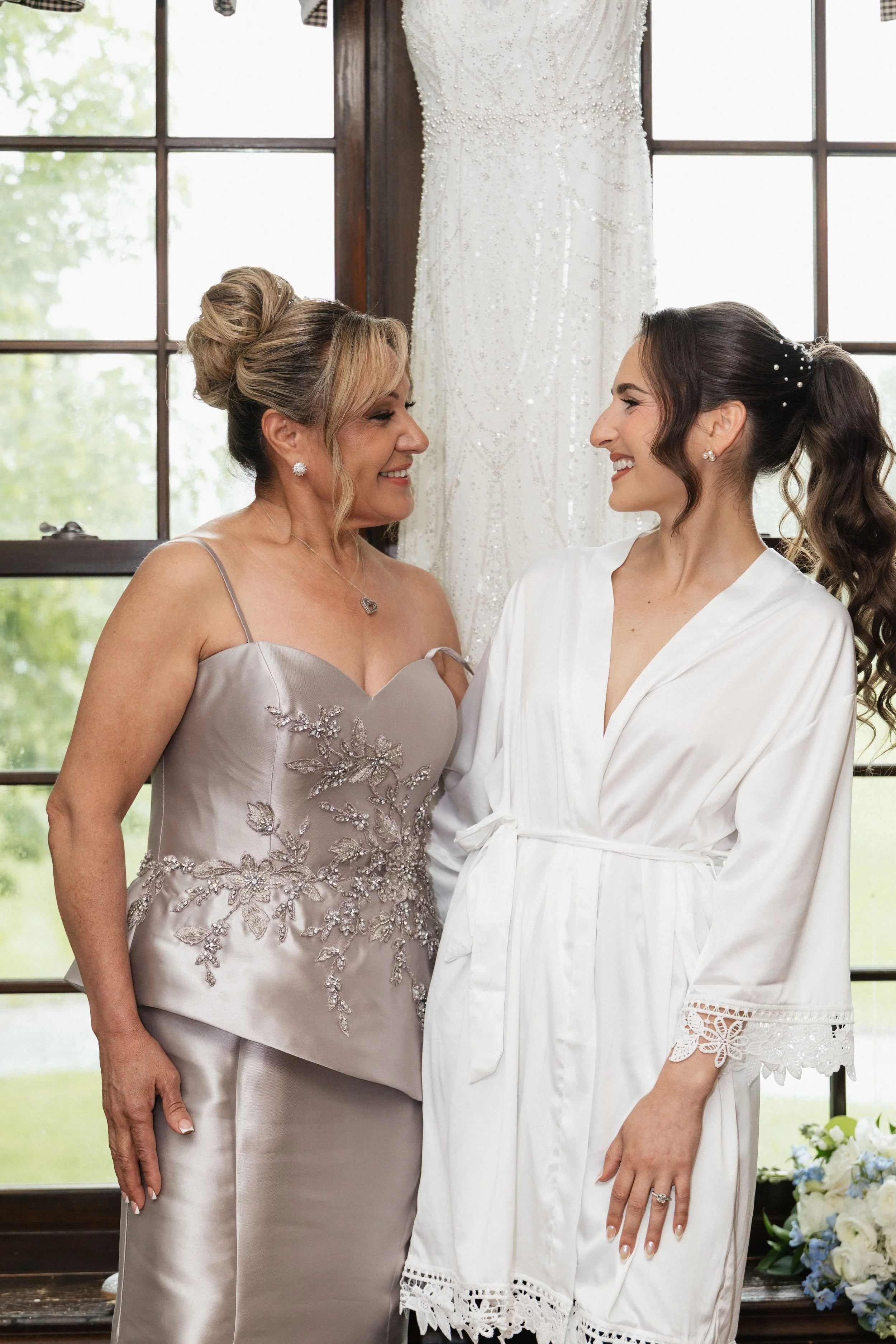 Two women, one older and one younger, smiling and facing each other indoors near large windows with a wedding dress hanging behind them. The older woman is wearing a light gray dress with floral embroidery, and the younger woman is wearing a white ro