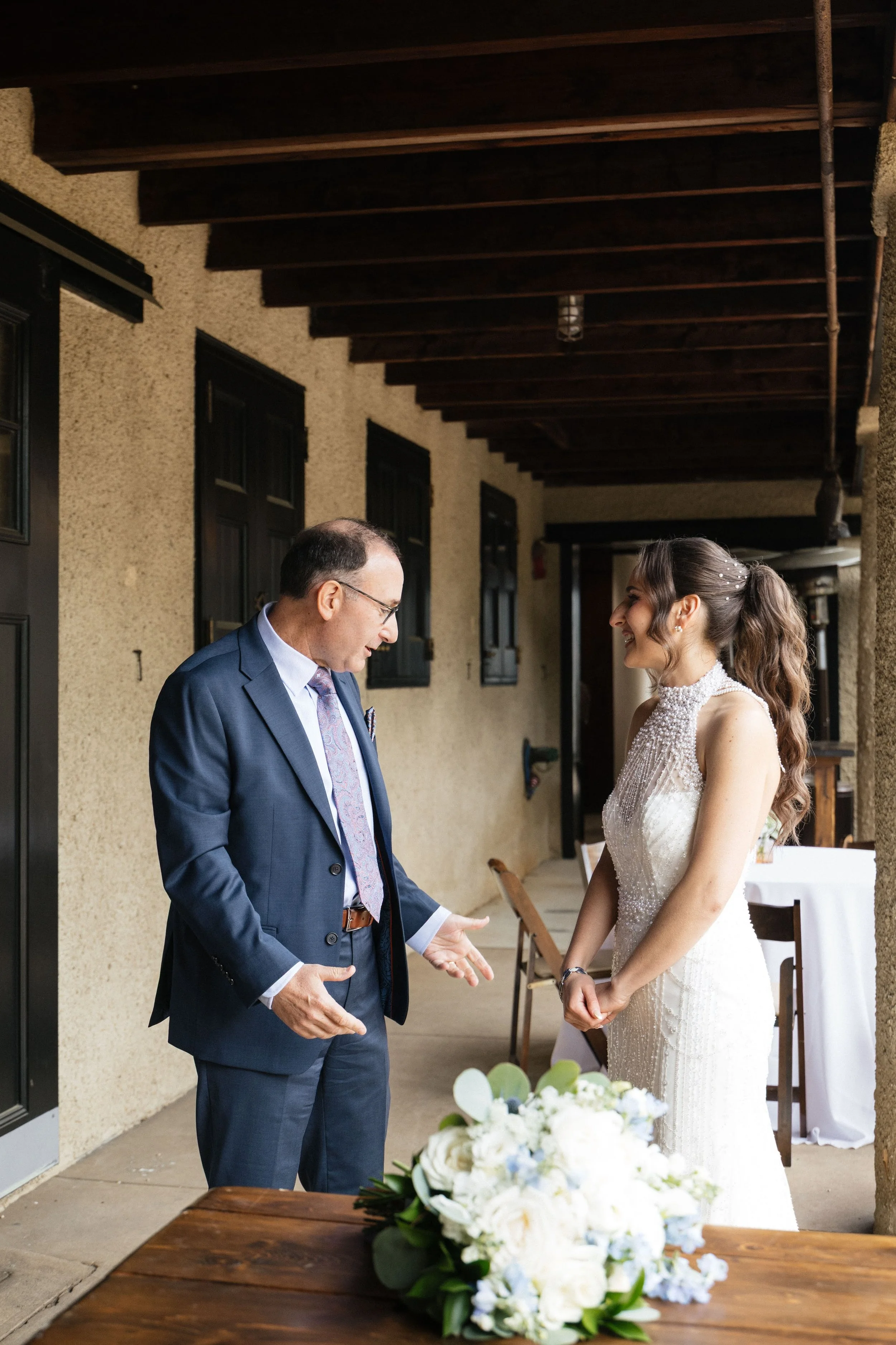 A man dressed in a suit and a woman in a wedding dress standing face-to-face, smiling, with the man talking and the woman listening, in an outdoor wedding reception setting.