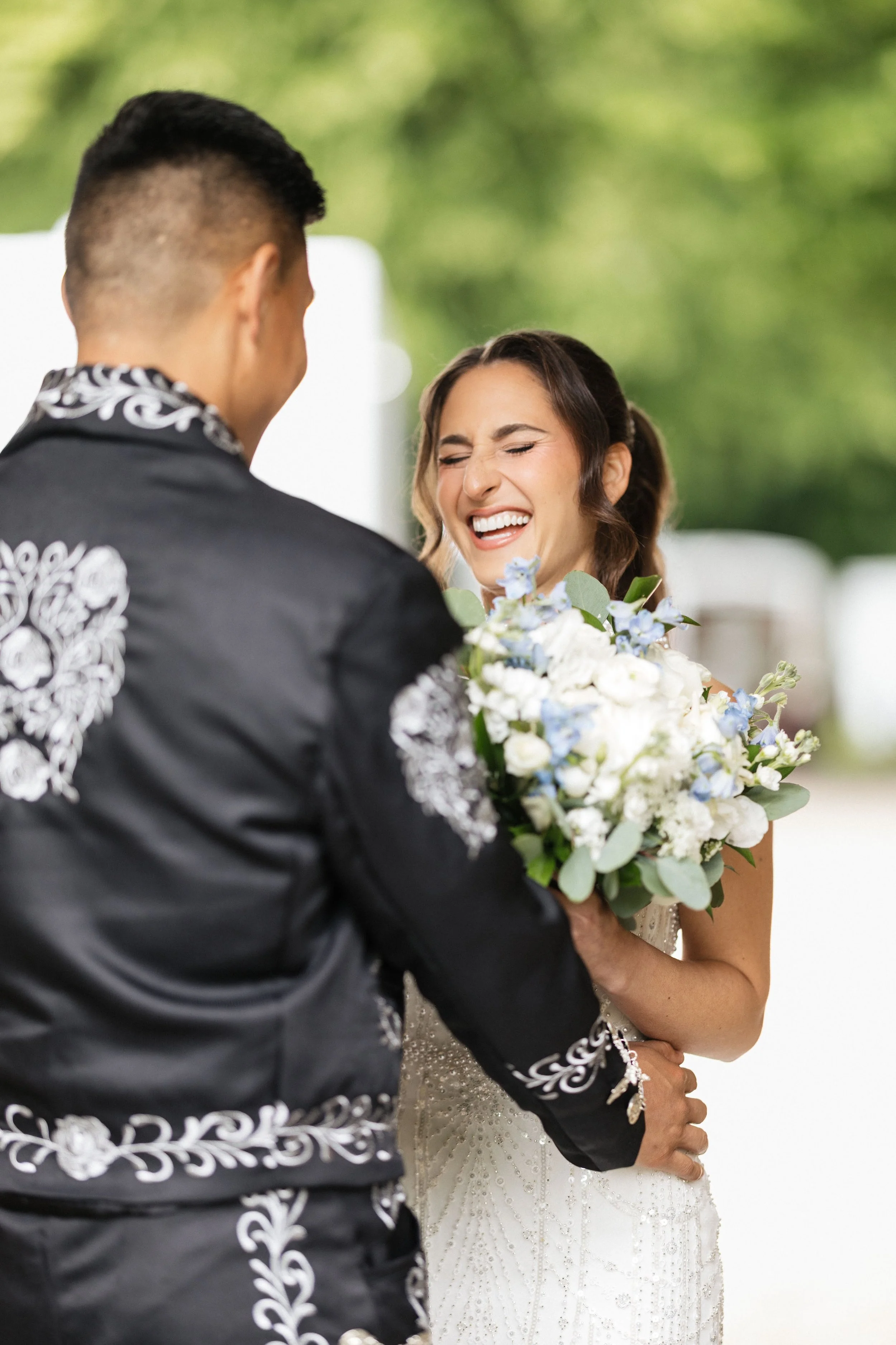 A bride and groom during their wedding ceremony, with the bride smiling and holding a bouquet of flowers, while the groom's face is not visible.