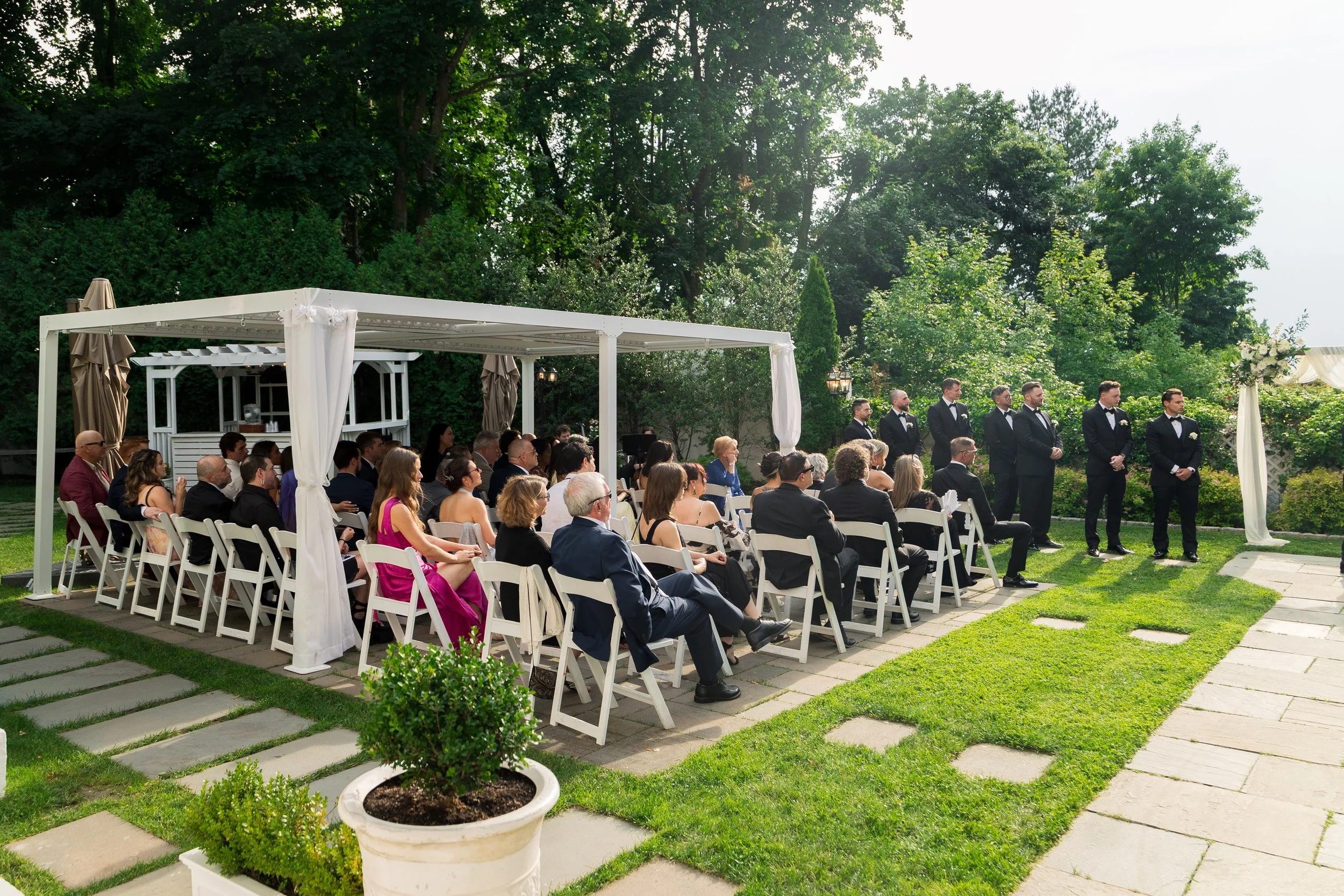 Outdoor wedding ceremony with guests seated on white chairs under a white canopy, surrounded by lush green trees and a grassy lawn.