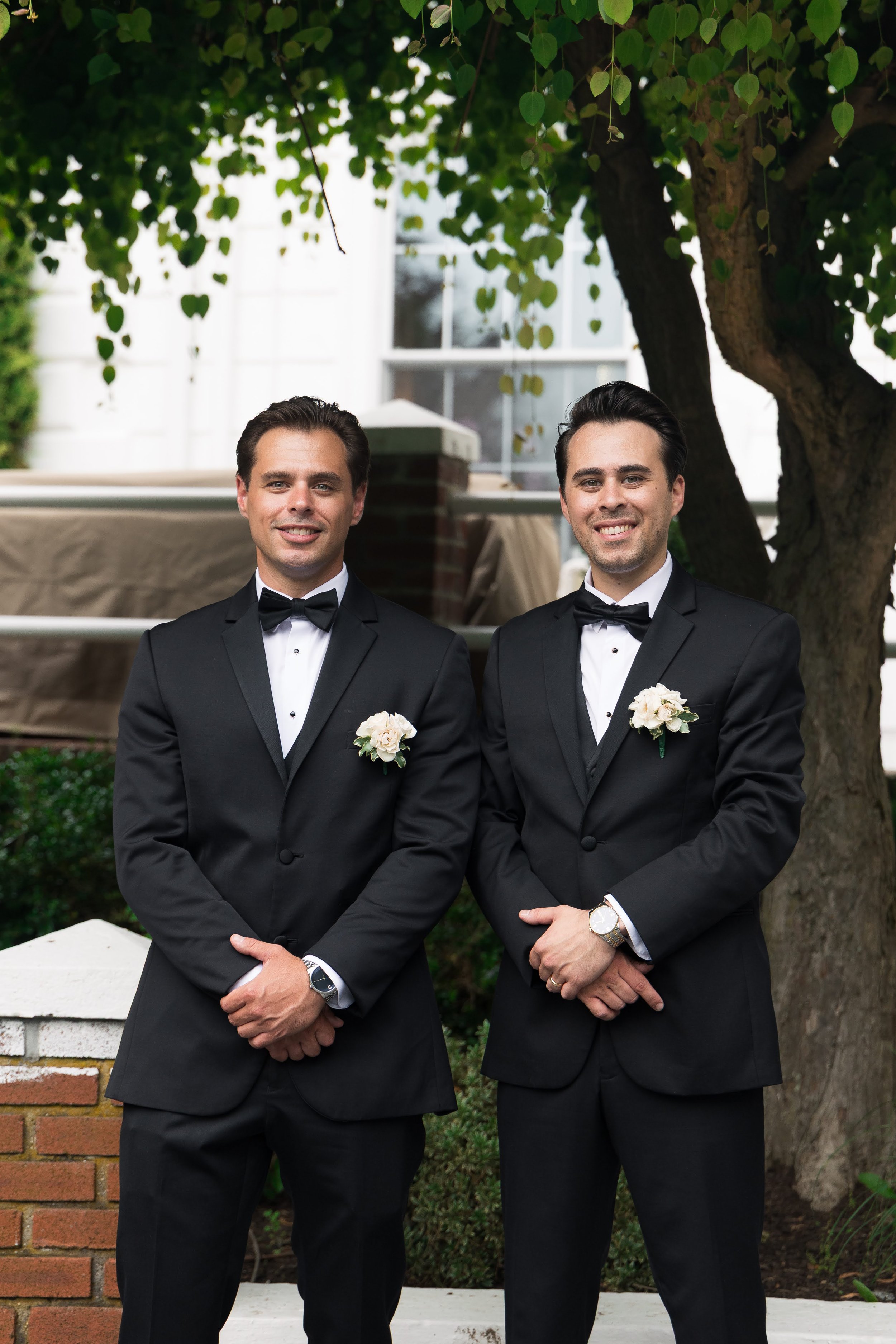 Two men in tuxedos with boutonnières standing outdoors under a tree, smiling at the camera.