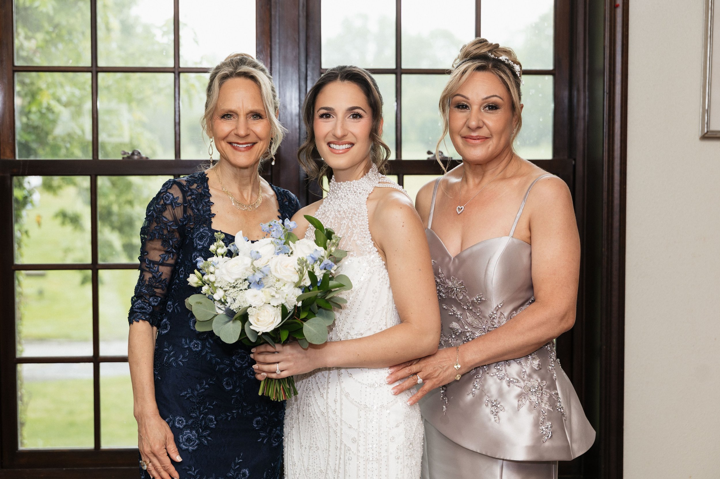 Three women, including a bride in a white wedding dress holding a bouquet, standing in front of a large window with greenery outside, smiling for a photo.