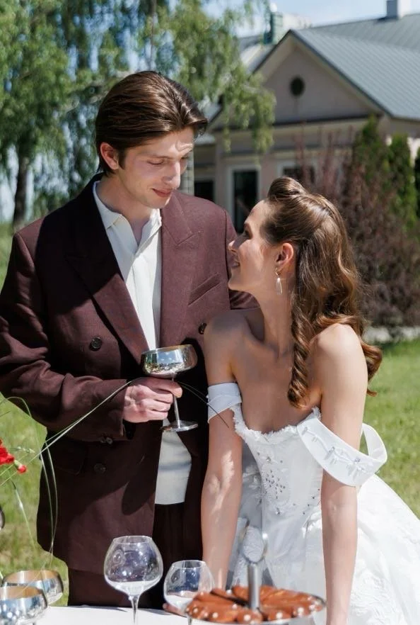 A man in a brown suit holding a glass of wine and a woman in a white dress sitting outdoors at a table, smiling at each other during daytime.