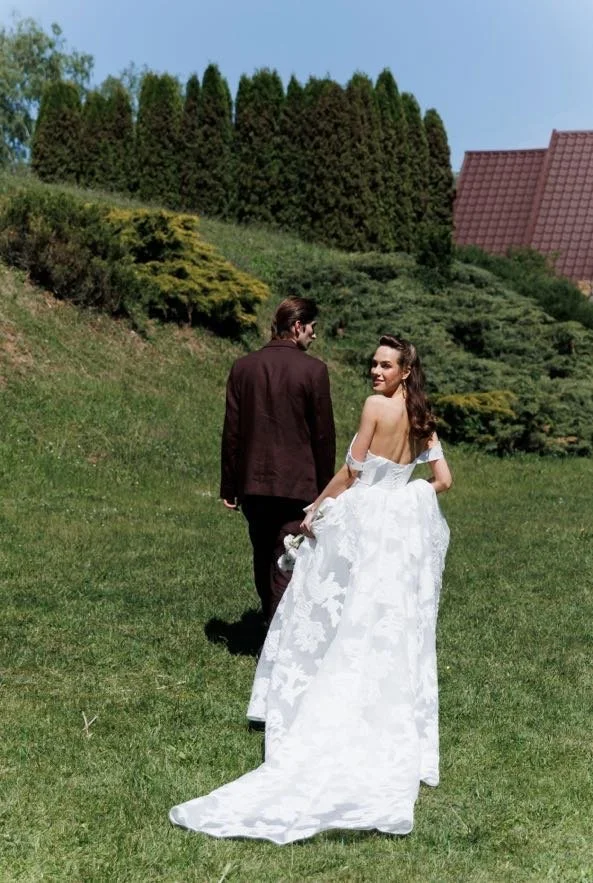 A bride and groom walking away on a grassy hill on a sunny day, with the bride looking back and smiling.