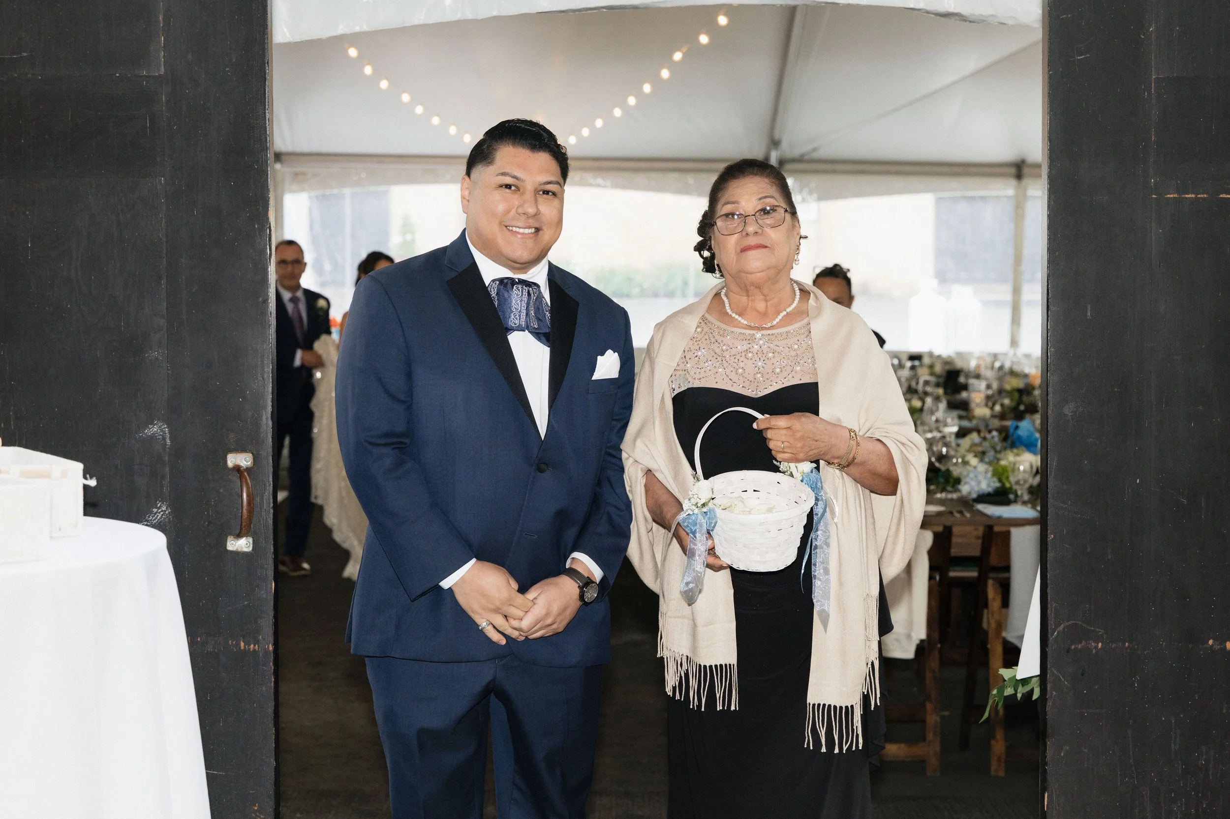 A young man in a blue tuxedo standing next to an older woman in a beige shawl holding a white basket at a wedding reception under a tent. Both are smiling, and there are people and tables with decorations in the background.