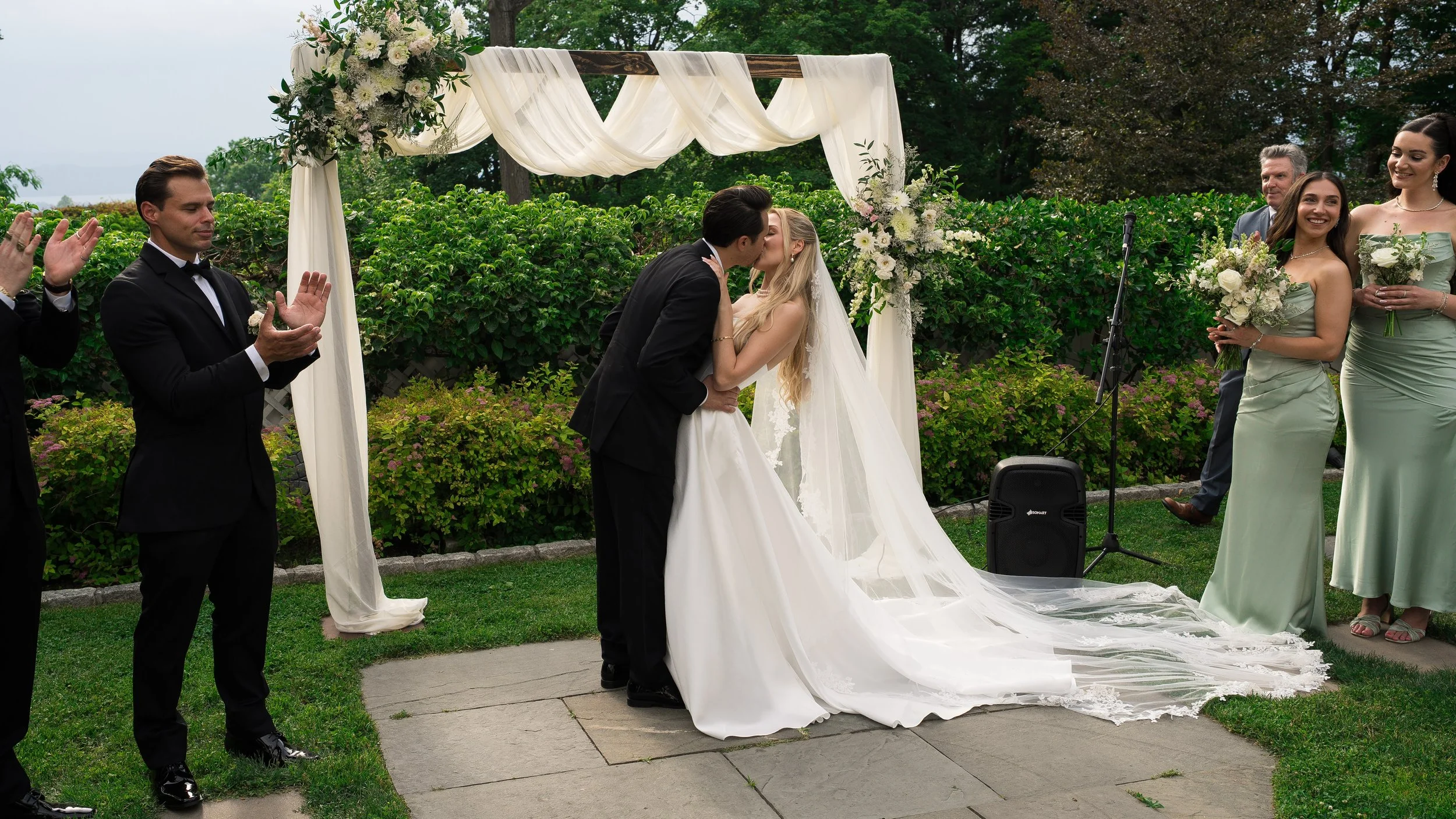 A couple is kissing during a wedding ceremony outdoors, surrounded by friends and family. The bride wears a white gown with a long train, and the groom wears a black tuxedo. There are floral decorations and greenery in the background.