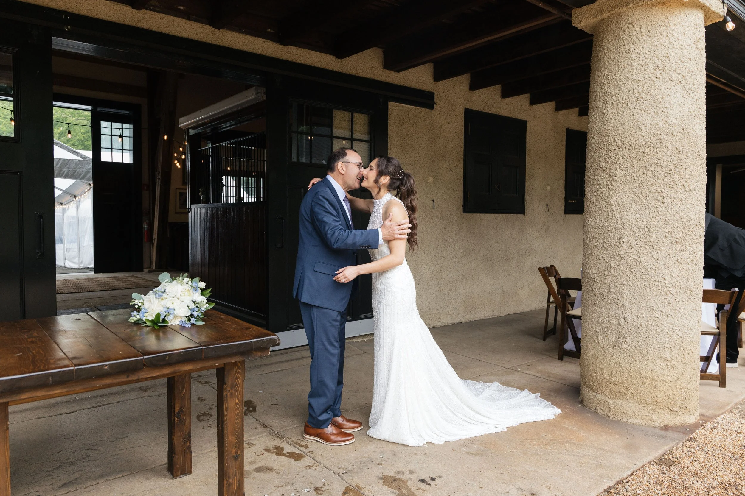A bride and groom smiling and embracing during a wedding ceremony indoors. The bride wears a white wedding gown with a train, and the groom wears a blue suit. There are flowers on a wooden table in the foreground, and rustic walls with black windows 