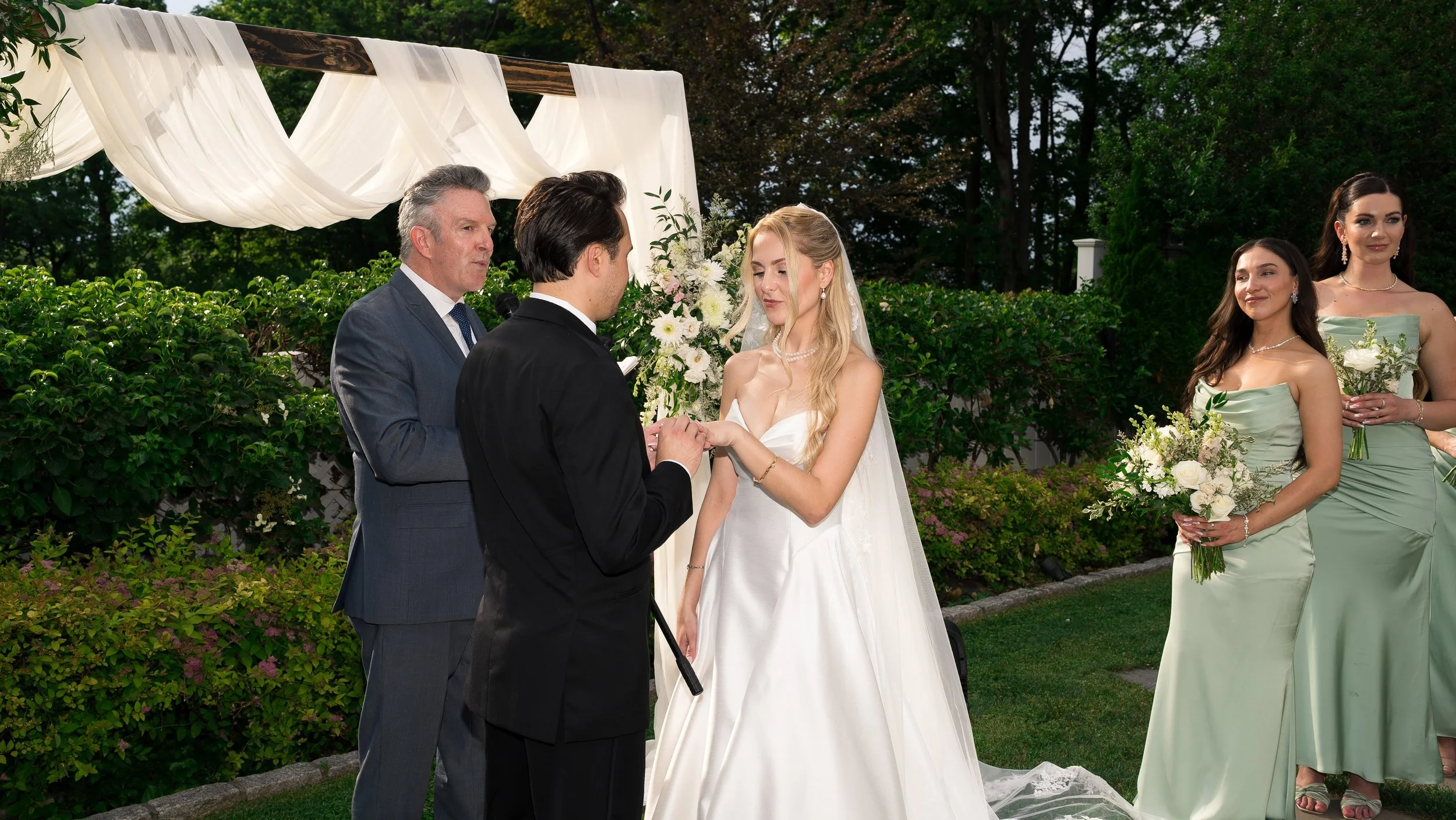 A bride and groom exchanging vows outdoors during a wedding ceremony, with a man officiating and bridesmaids holding bouquets in the background.