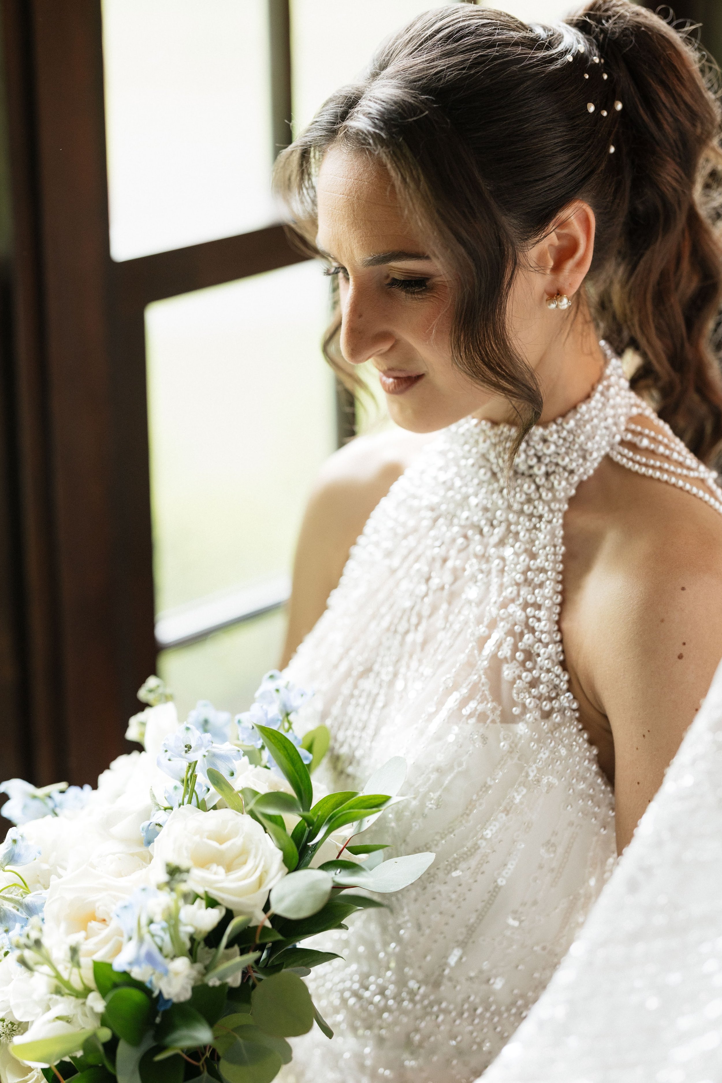 A bride in a beaded wedding dress holding a bouquet of white roses and greenery, standing near a window.